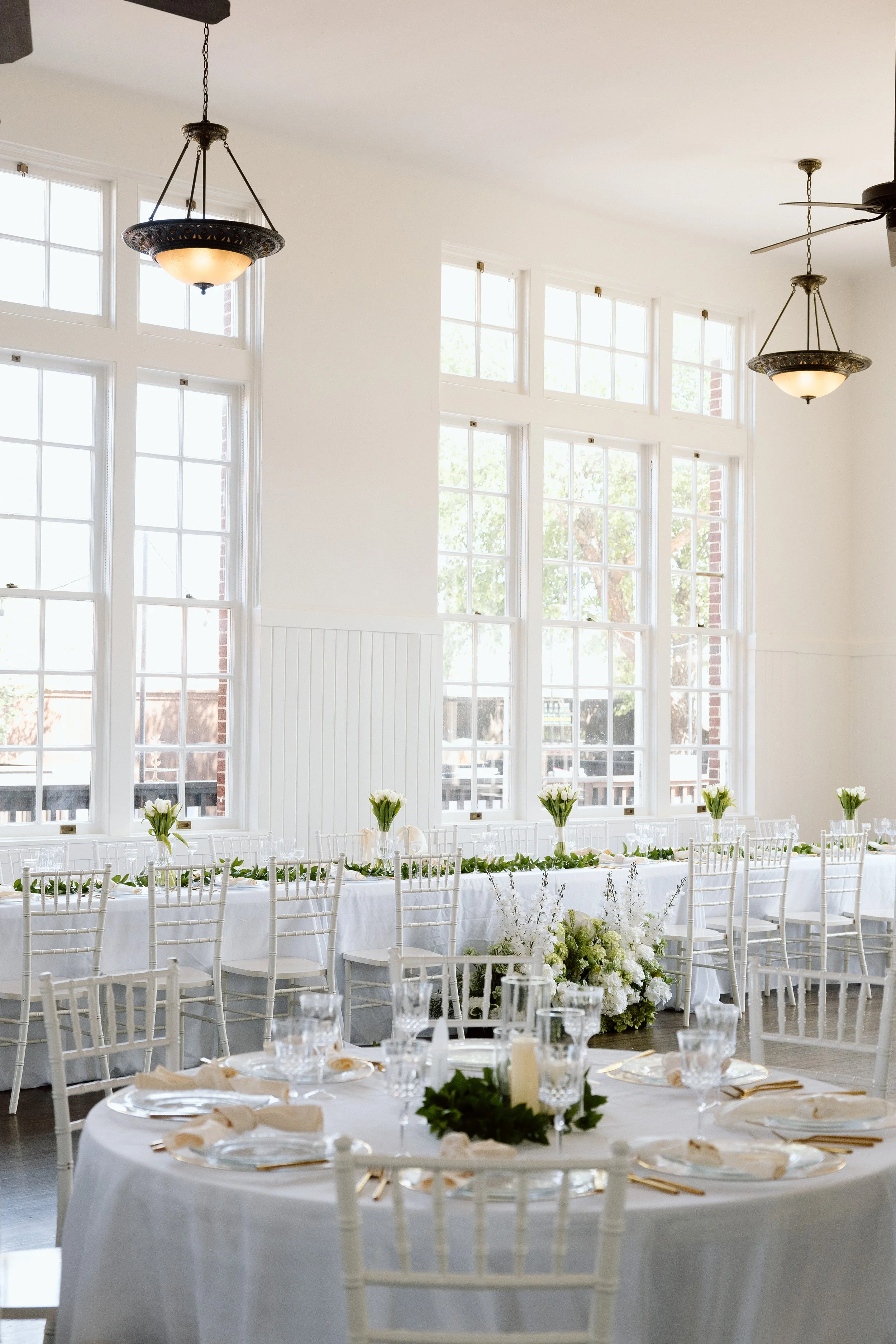 Elegant indoor wedding reception setup with round tables covered in white tablecloths, decorated with white flowers, greenery, candles, and gold cutlery. The room features large windows allowing natural light and hanging light fixtures.