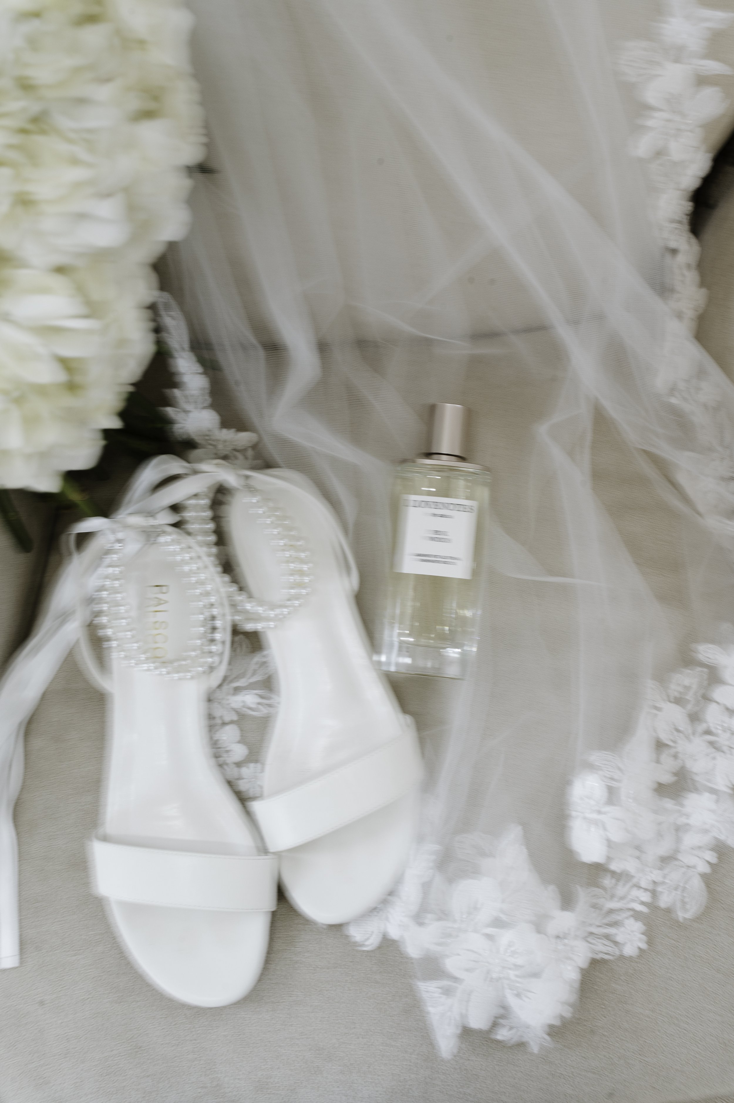 White wedding shoes on a display stand, with white flowers and a bottle of perfume nearby on a lace fabric background.