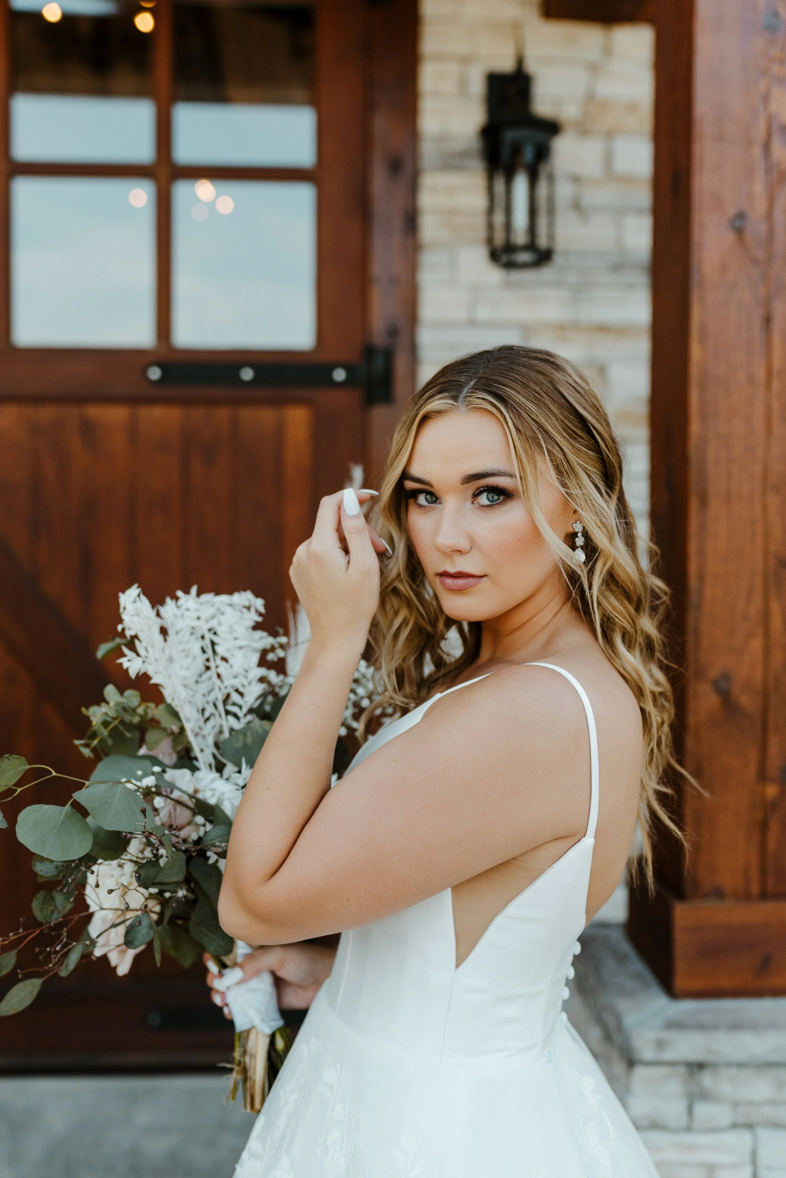 A woman in a white wedding dress holding a bouquet, standing outdoors near a rustic wooden building with a window and a lantern, with greenery around her.