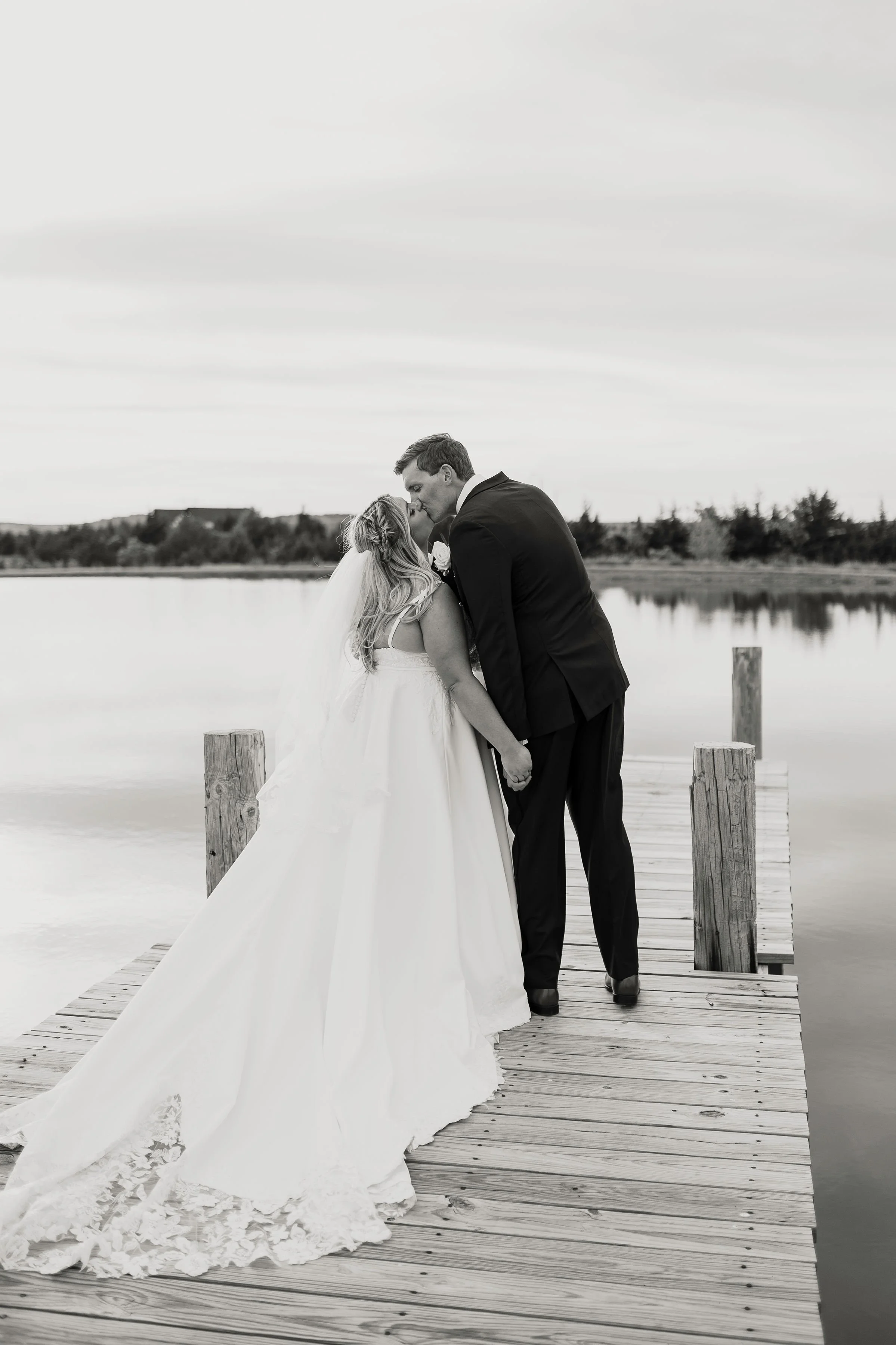 Black and white photo of a bride and groom kissing on a wooden dock near a body of water, with trees in the background.