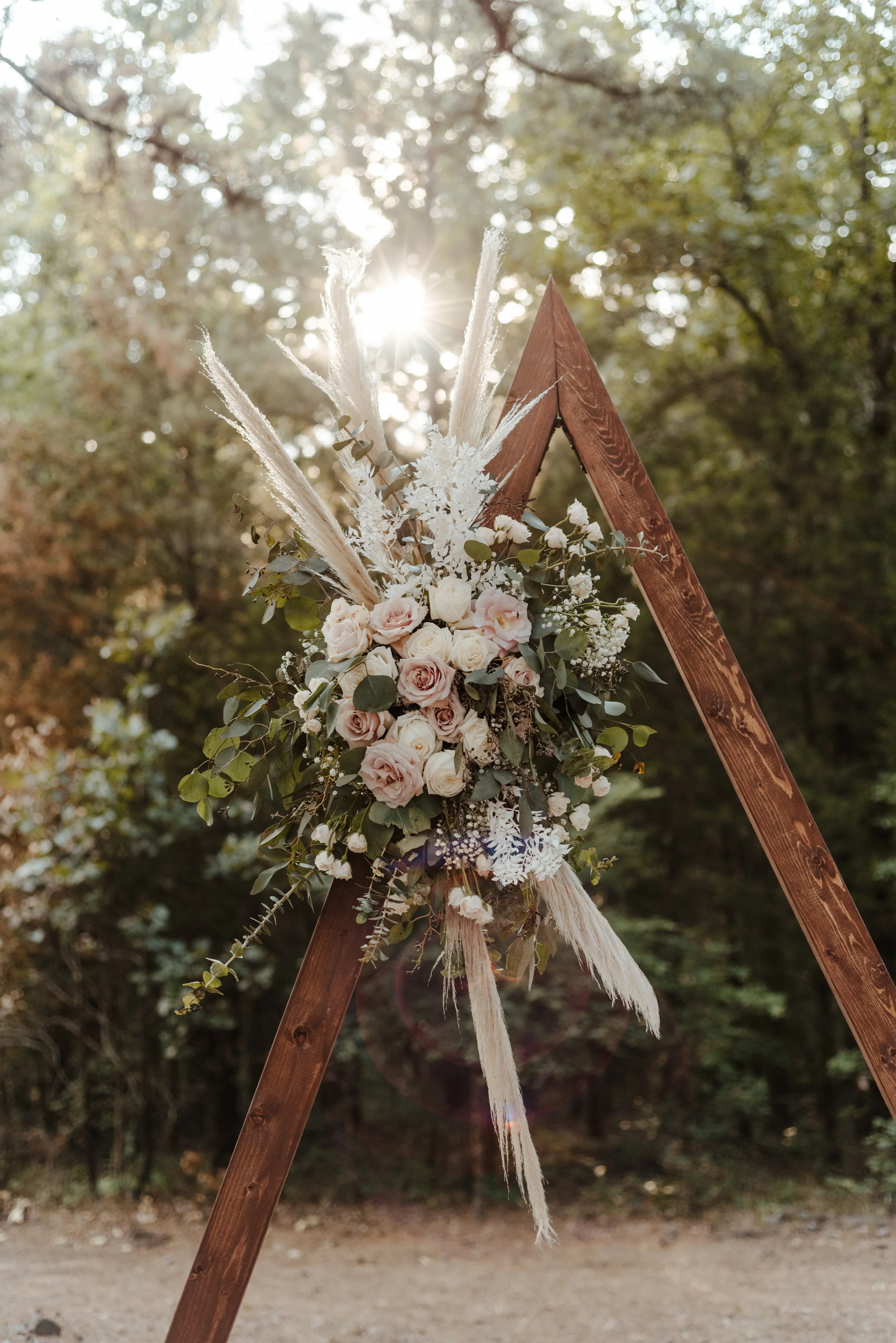 A wooden triangular structure decorated with white and blush pink roses, greenery, and dried pampas grass, set outdoors with sunlight filtering through the trees.