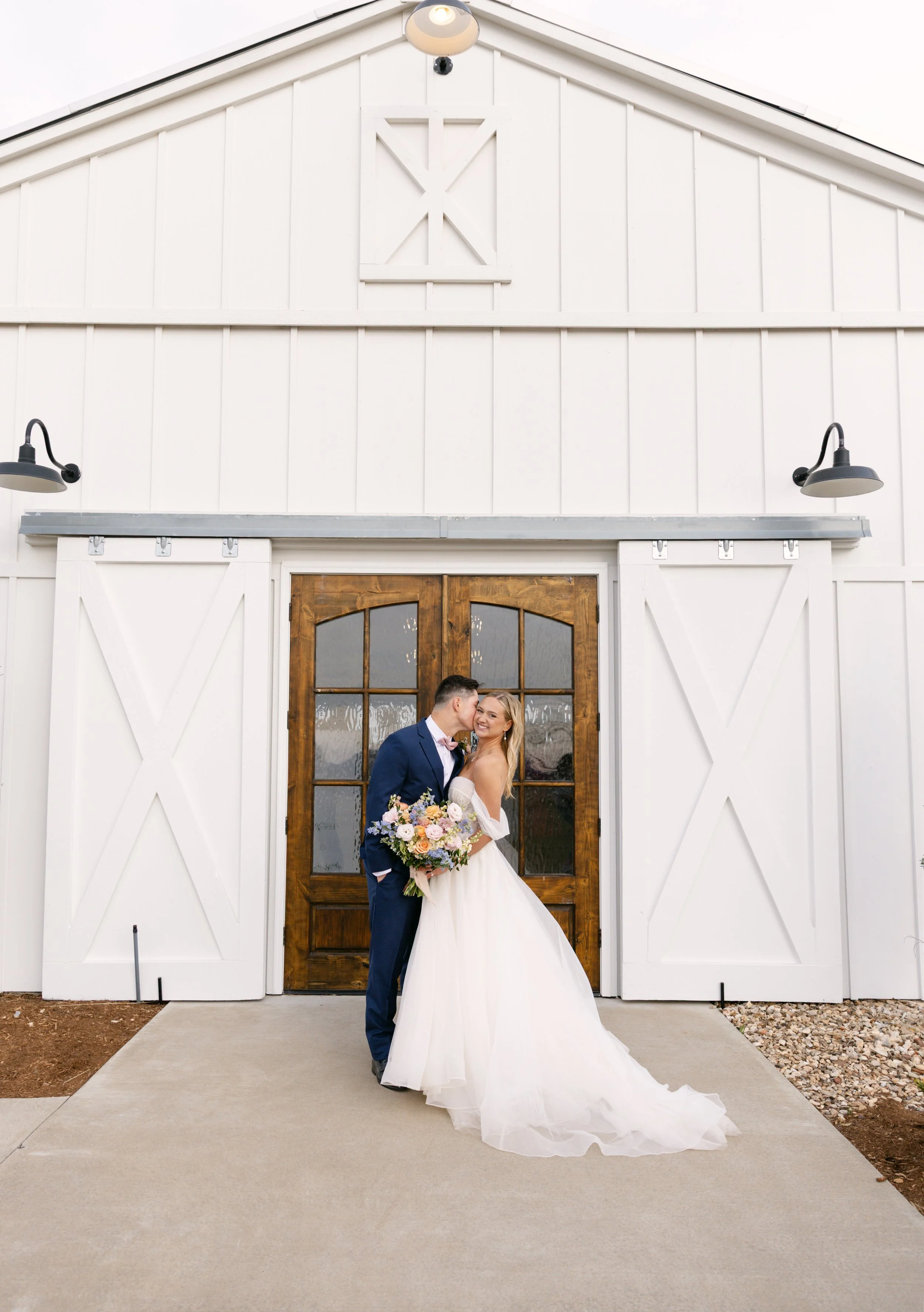 A couple dressed in wedding attire standing in front of a white barn with wooden doors, holding a bouquet of flowers, and sharing an intimate moment.