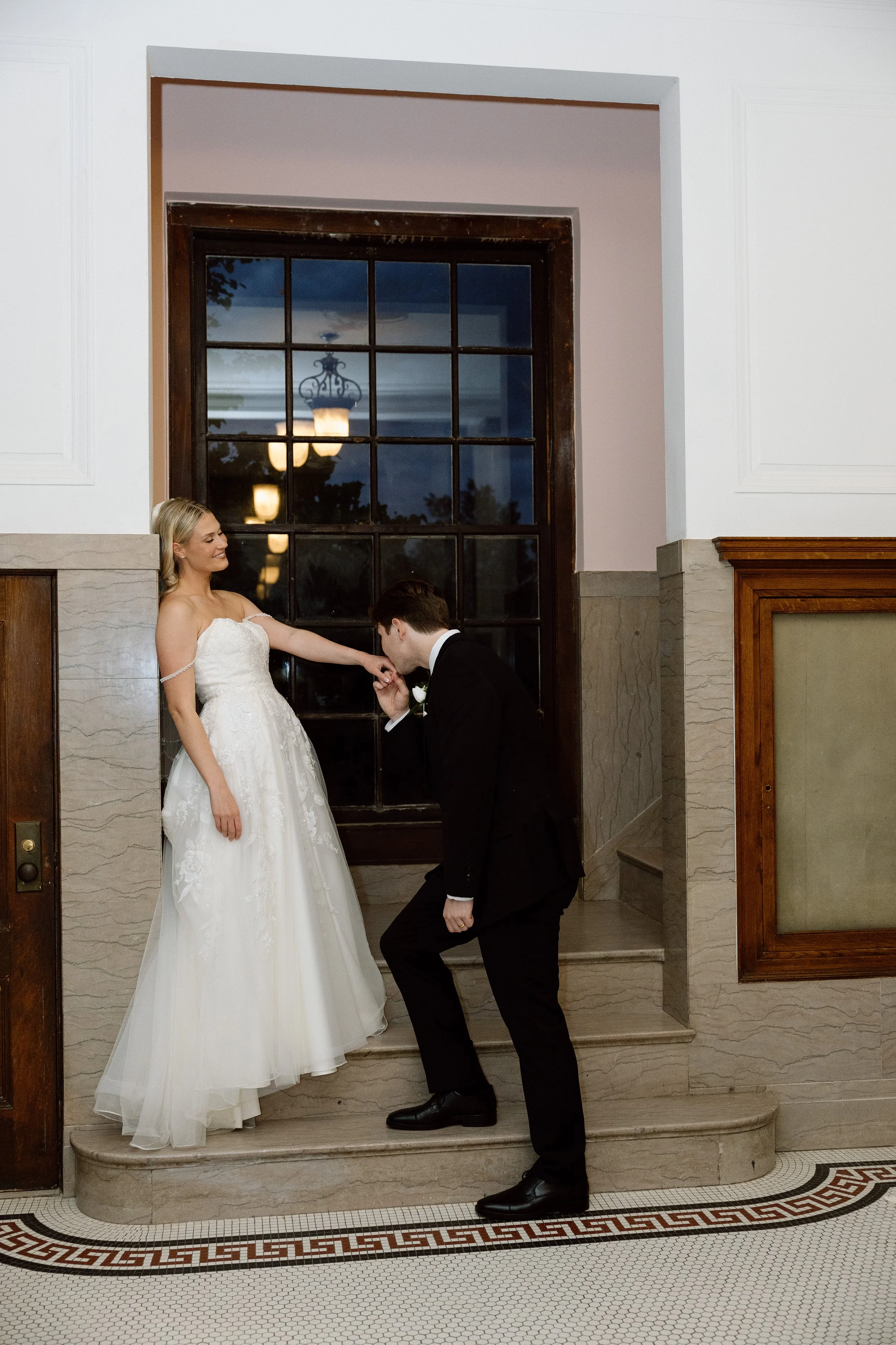 A bride in a white wedding dress is standing on steps, holding hands with a groom in a black suit who is kneeling and kissing her hand inside a building with large windows and vintage light fixtures reflected in the glass.