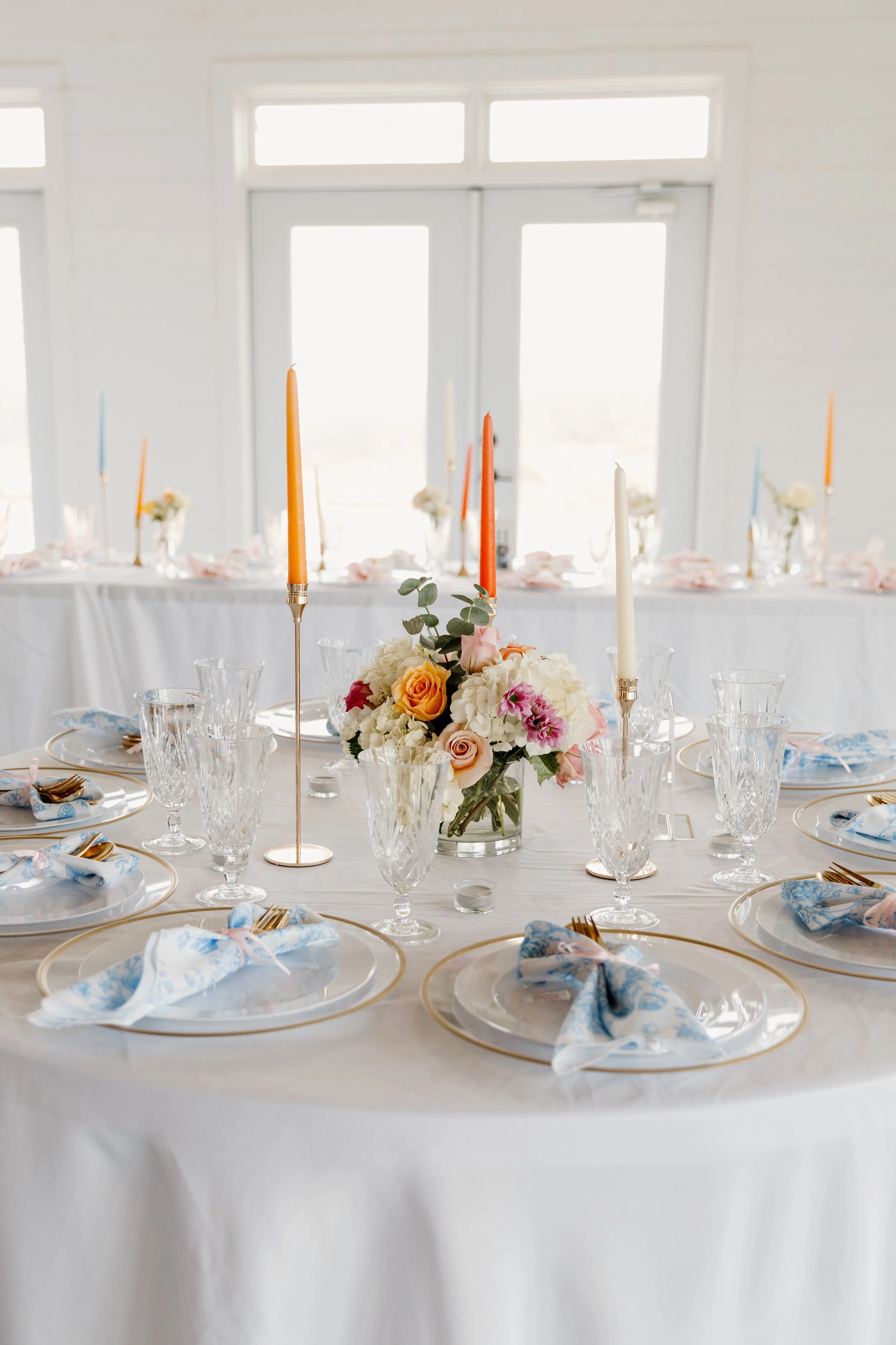 Elegant round dining table set for a formal event with white tablecloth, floral centerpiece, tall multicolored candles, crystal glassware, and blue patterned napkins on white plates with gold rims, in a bright room with large windows.