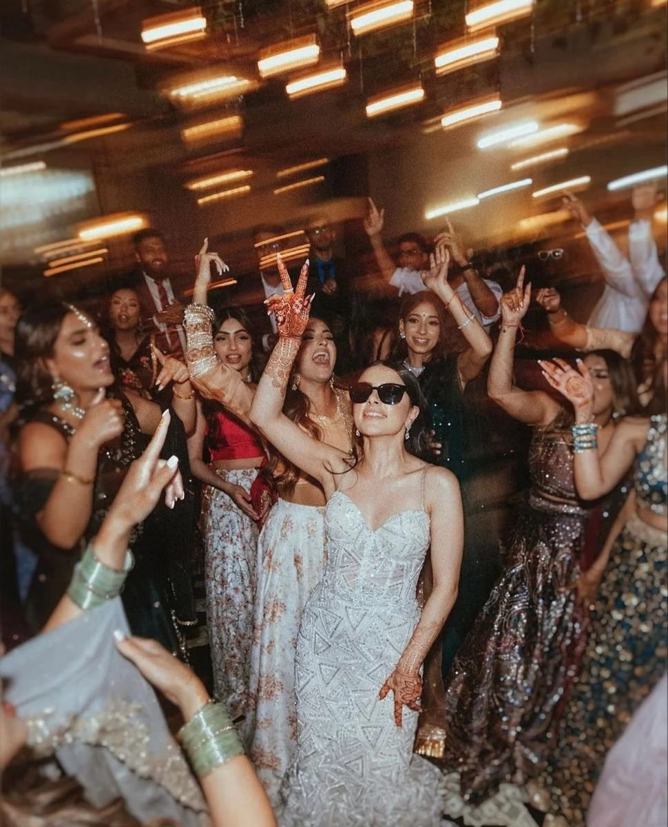 Group of women dancing at a festive wedding reception, with the bride in a white embellished dress and sunglasses in the foreground