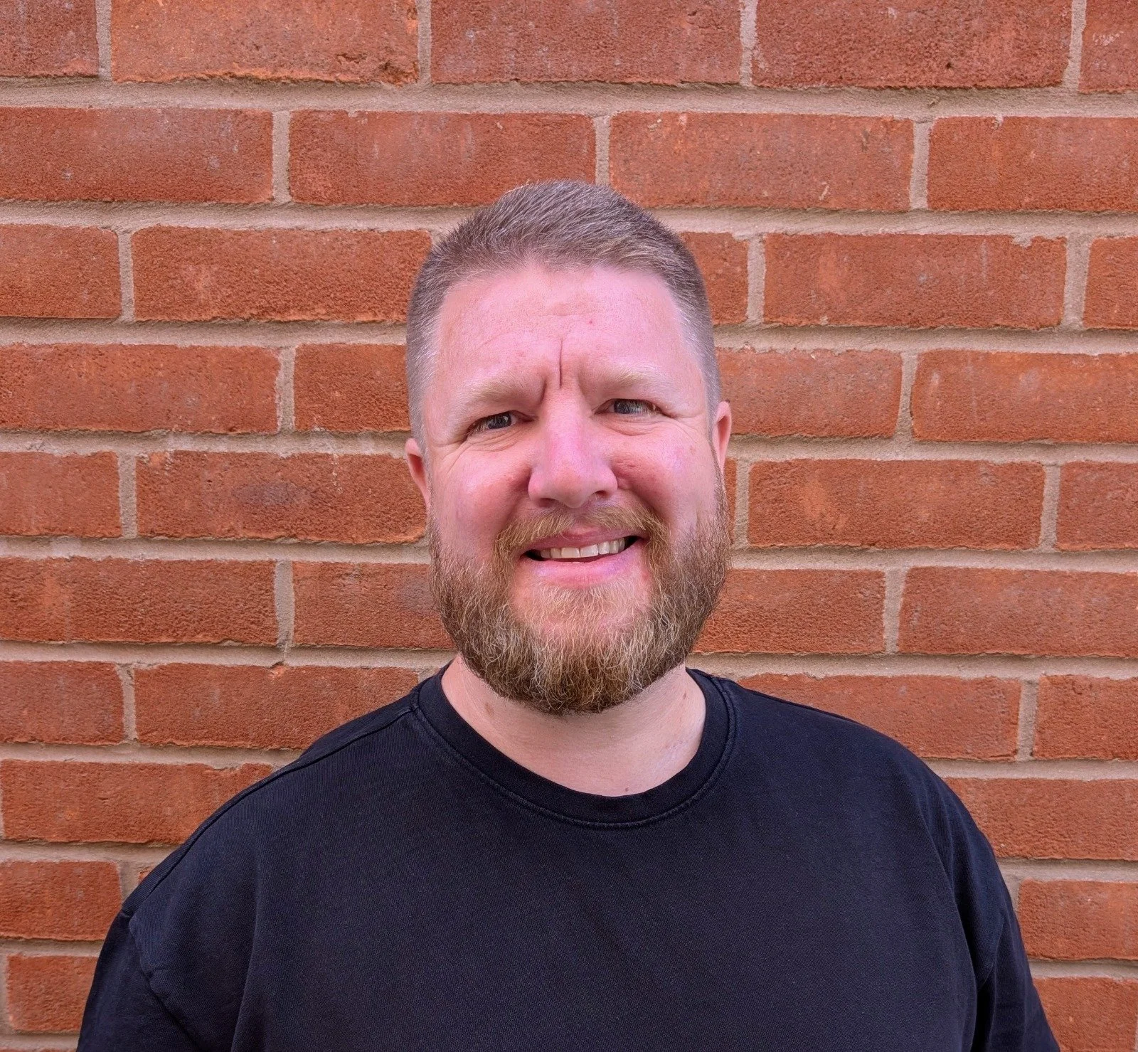 A man with a beard and short hair smiling in front of a brick wall, wearing a black shirt.