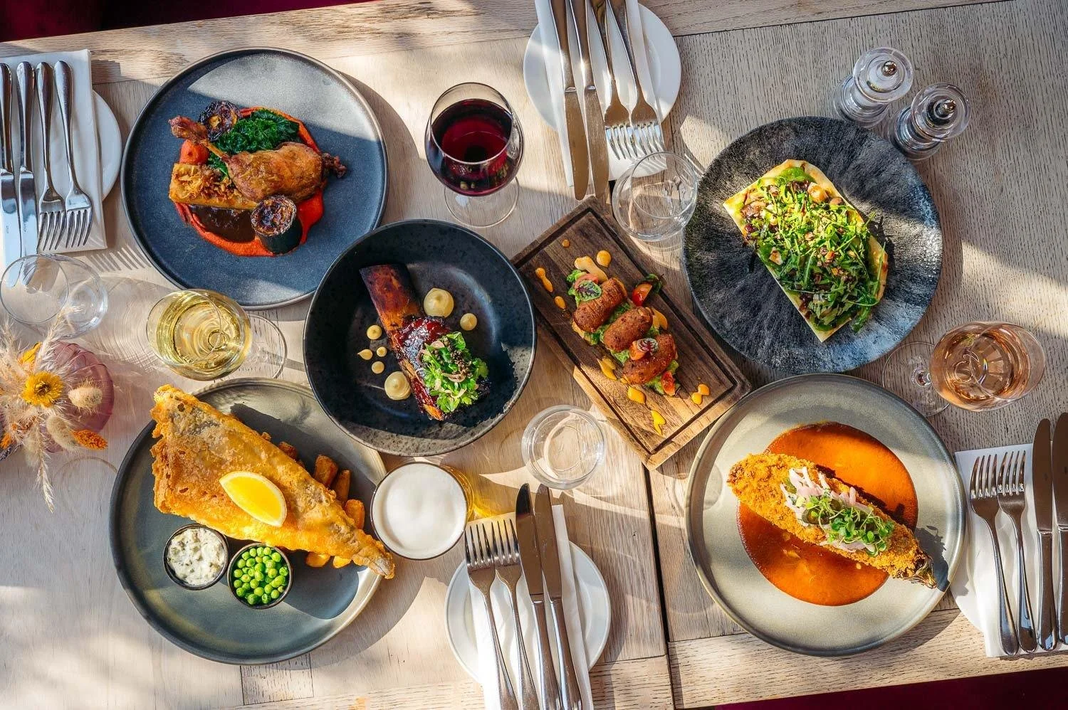 A table set with various dishes including a roasted chicken with vegetables, a slice of pie with lemon on top, a plate of croquettes with sauce and garnishes, a dish with greens on a rectangular plate, and a fillet of fish in a red sauce, along with glasses of wine and water, cutlery, and small sauce containers.