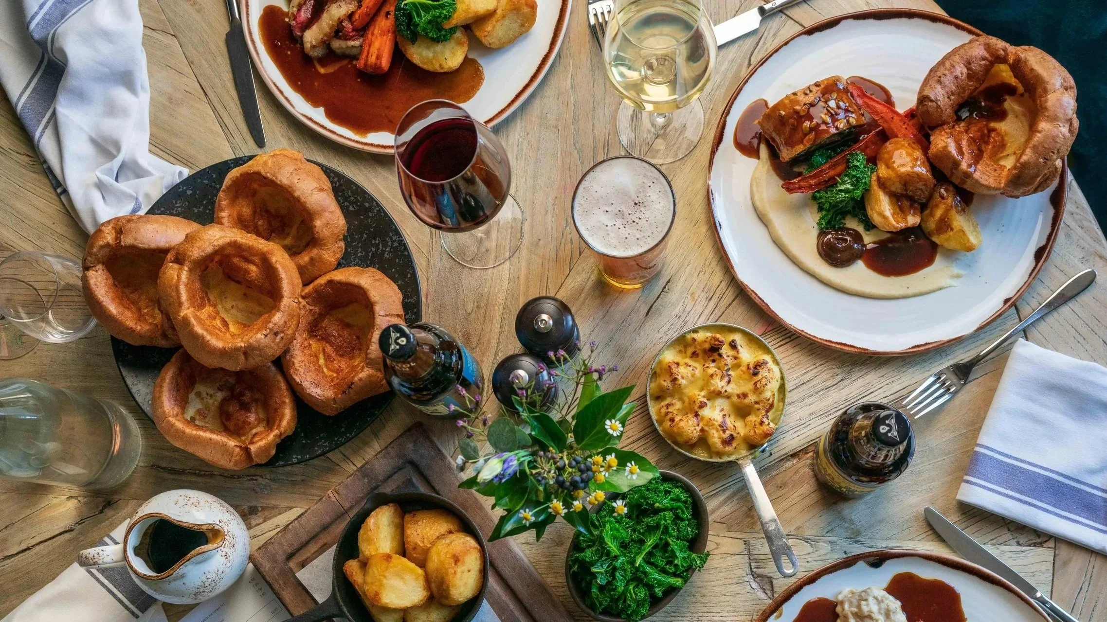 A table set with various dishes, including large Yorkshire puddings, roasted vegetables, mashed potatoes, a bowl of cheese, and glasses of red and white wine, beer, and a beer bottle, with cutlery and napkins arranged around.