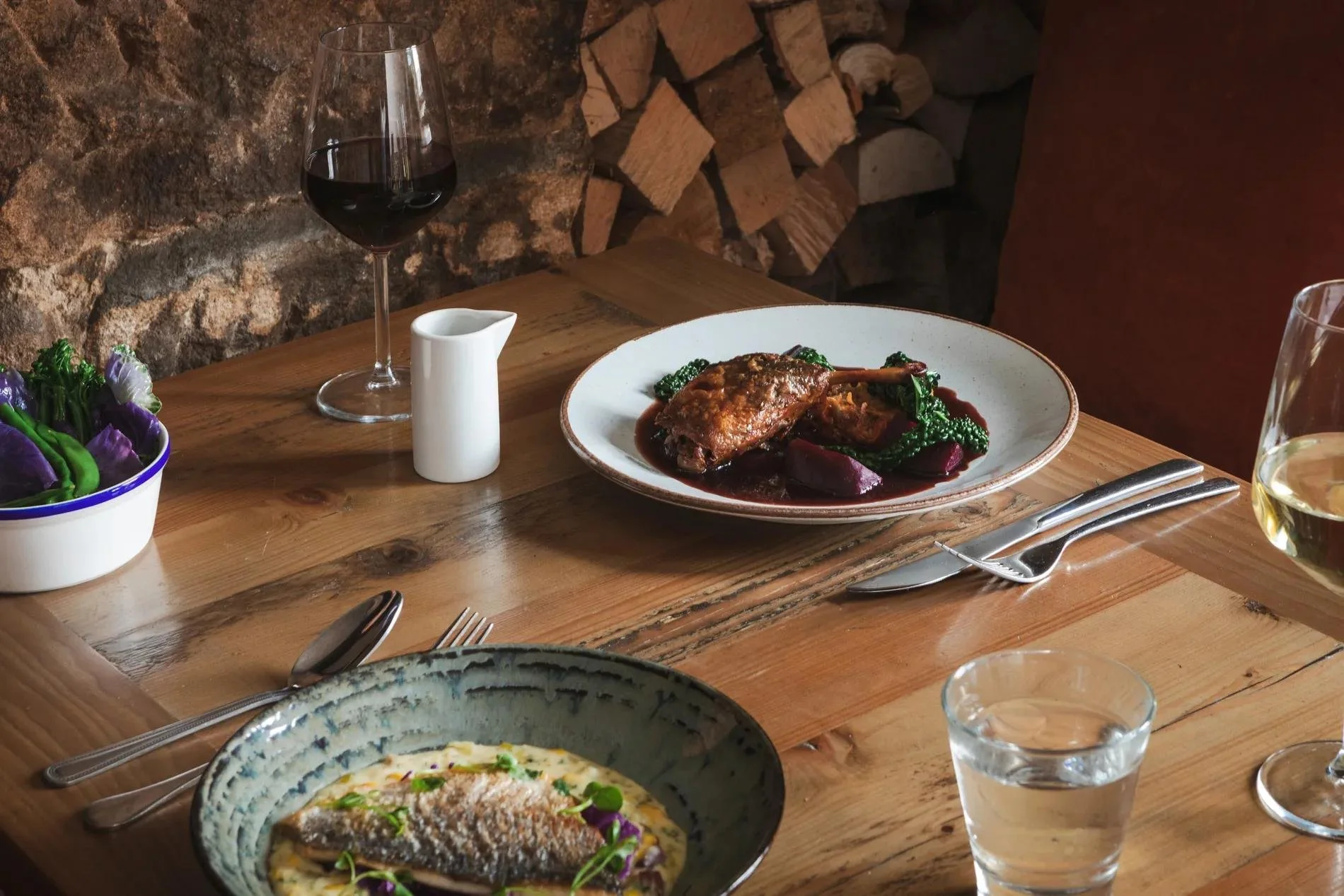 A wooden table set with a glass of red wine, a white ceramic creamer, a bowl of colorful vegetables, a plate with roasted meat, cooked greens, and roasted vegetables, and a glass of white wine, with a rock wall and stacked firewood in the background.
