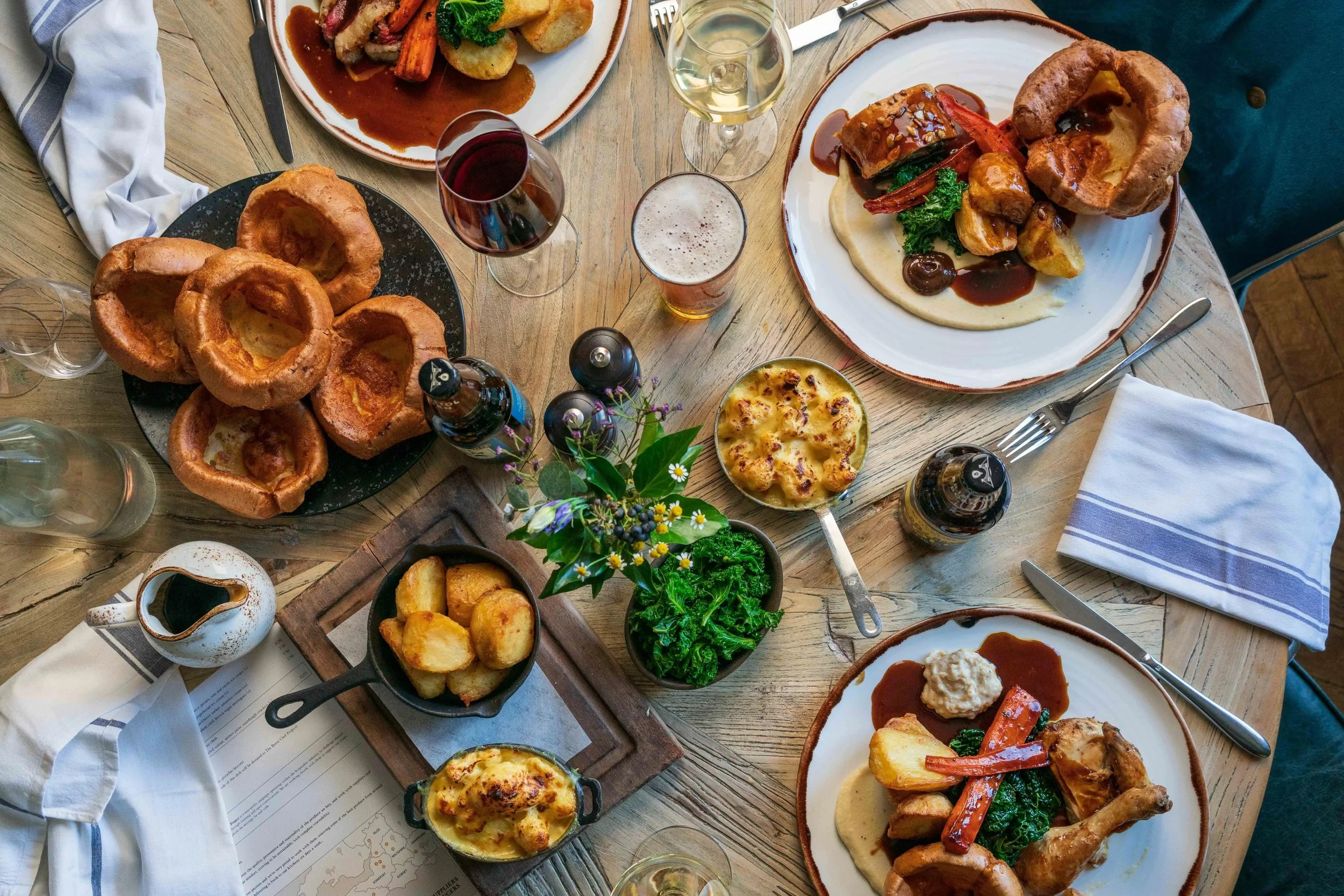 A table set with various plates of food including Yorkshire pudding, roasted vegetables with sauce, mashed potatoes, and salads. There are glasses of red and white wine, beer, water, and a small flower arrangement on the table. Napkins and cutlery are also visible.