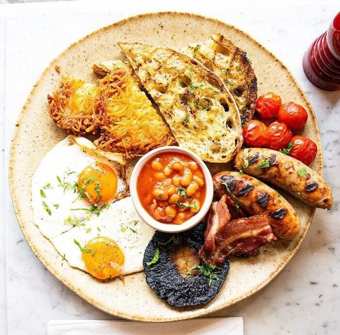 A breakfast plate with fried eggs, grilled flatbread, roasted cherry tomatoes, beans, bacon, sausages, and black pudding, served with ketchup.
