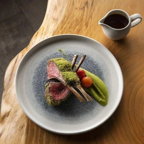 A plate with a pinkish-red piece of meat, garnished with green powder, accompanied by green puree and two cherry tomatoes, with a small white pitcher of dark sauce on the side on a wooden table.
