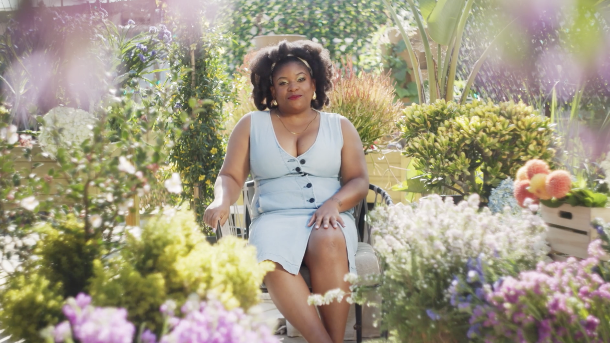 A woman sitting on a bench in a garden filled with colorful flowers and greenery, smiling at the camera.