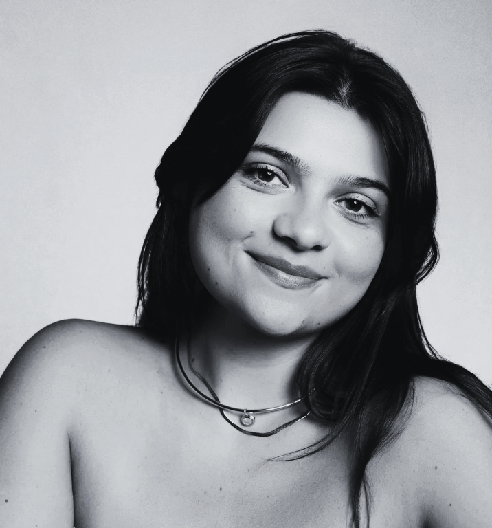 Black and white portrait of a young woman with long dark hair, smiling softly, facing the camera with bare shoulders and layered necklaces.