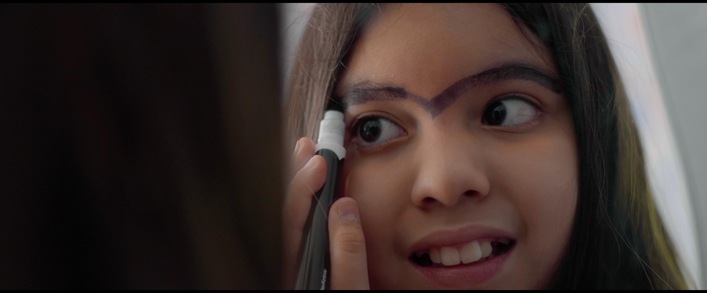A young woman with dark hair and eyebrows is drawing on her face with a black eyeliner, creating a thick, angular line across her forehead and over her eyebrows.