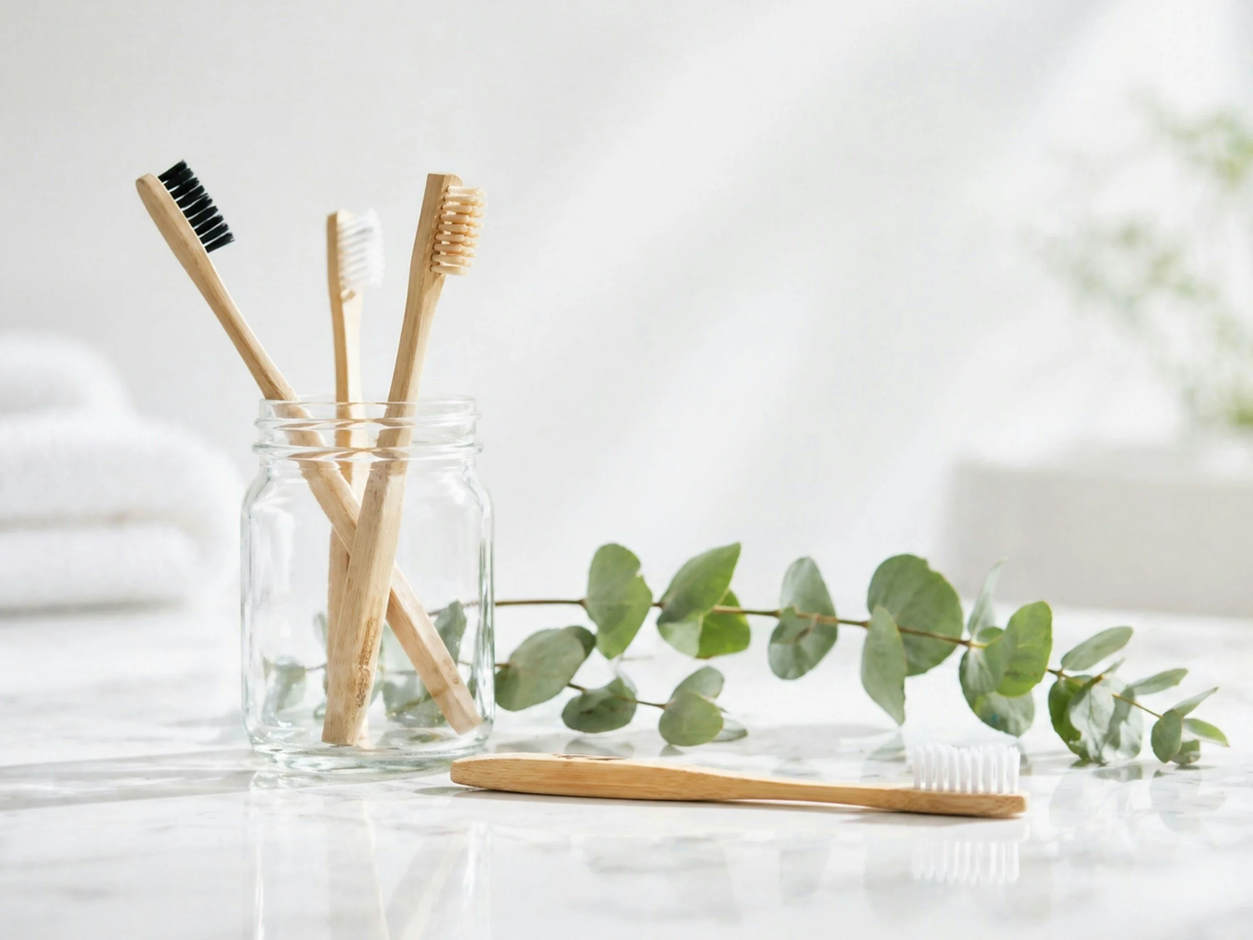 Three bamboo toothbrushes in a clear glass jar, with eucalyptus leaves and a wooden toothbrush lying flat on a white surface, against a soft white background.