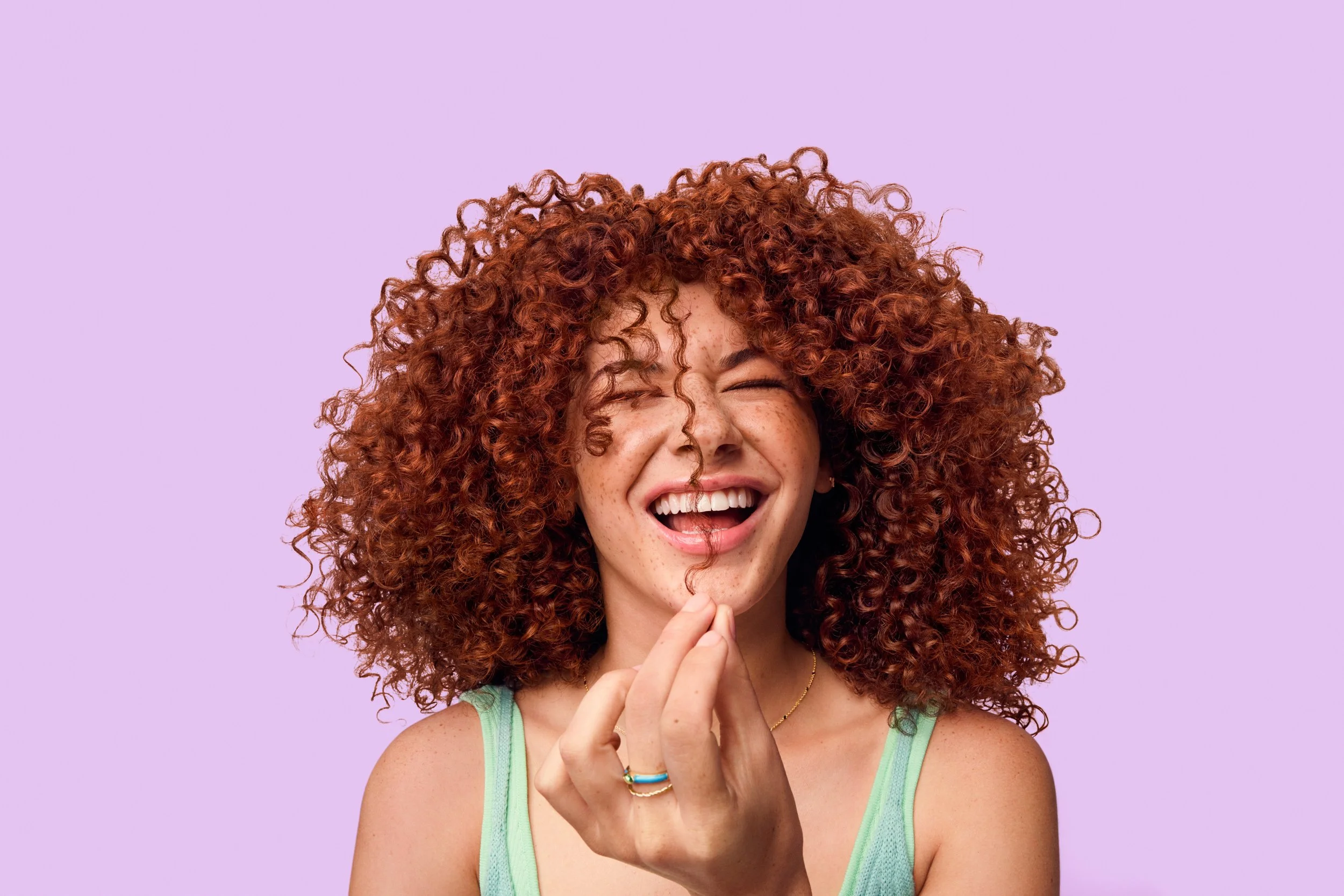 Freckled woman with voluminous curly red hair laughing against a lavender background