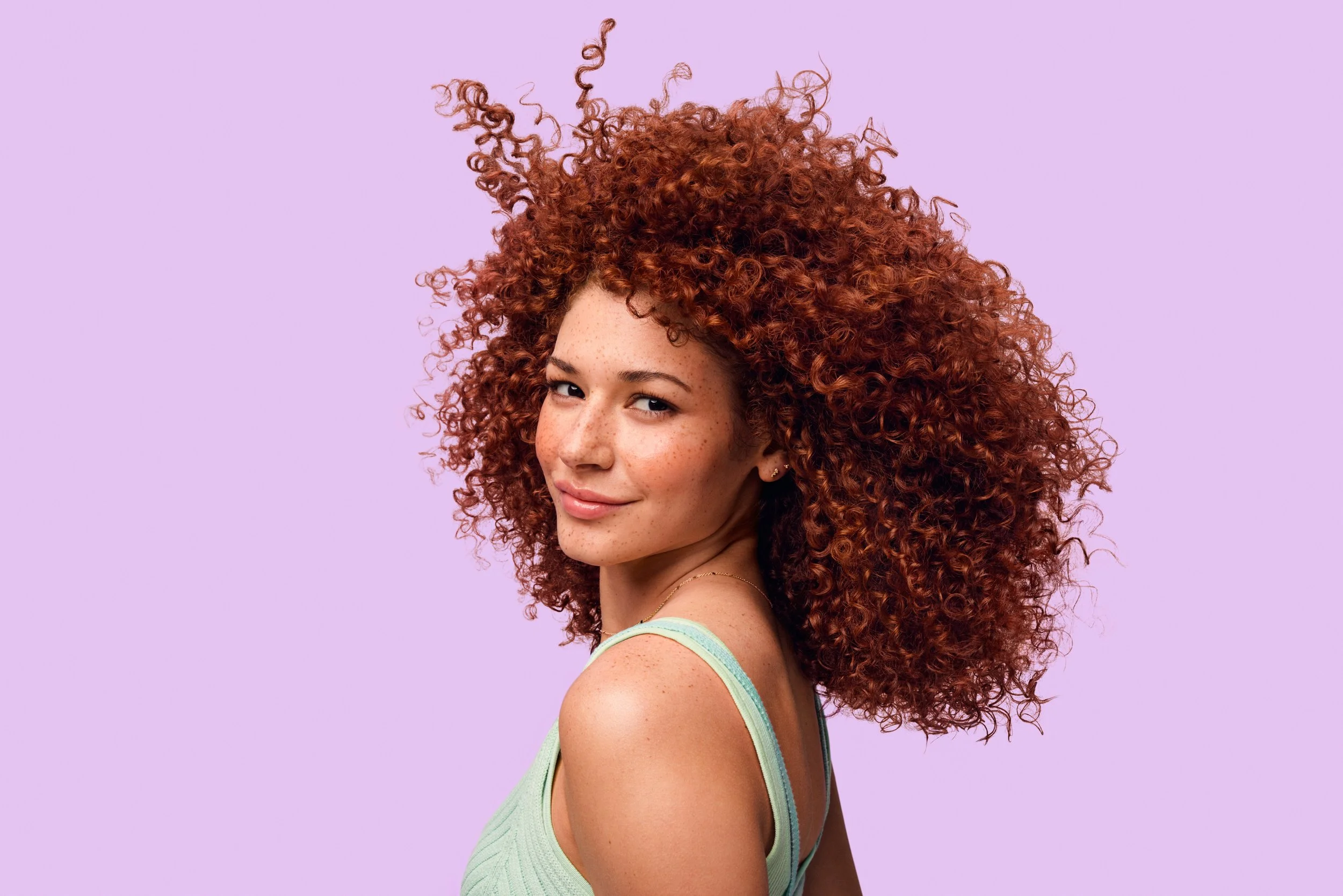 Portrait of a smiling woman with voluminous curly red hair against a lavender background