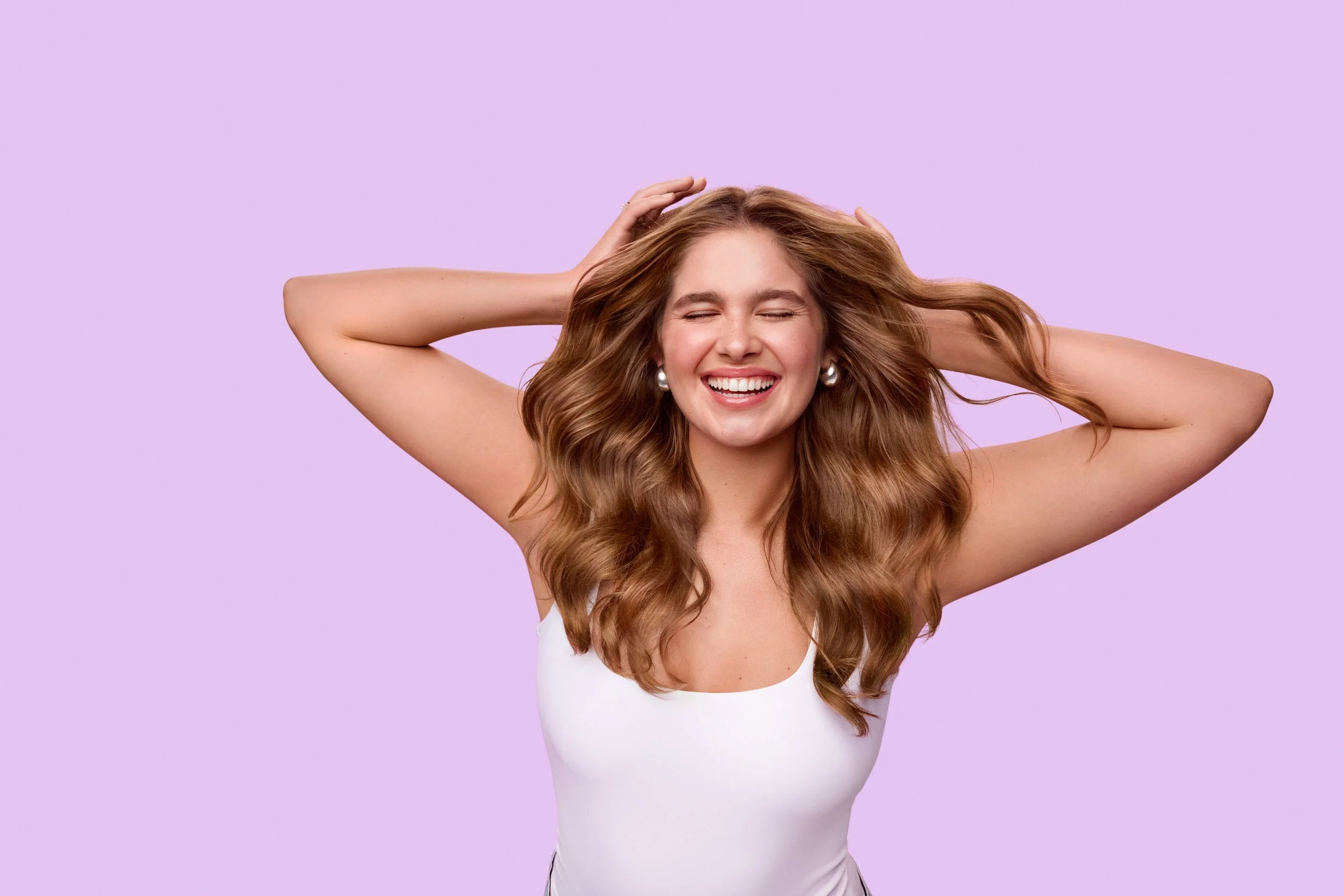 Smiling woman with long wavy brown hair posing with her hands behind her head against a lavender background