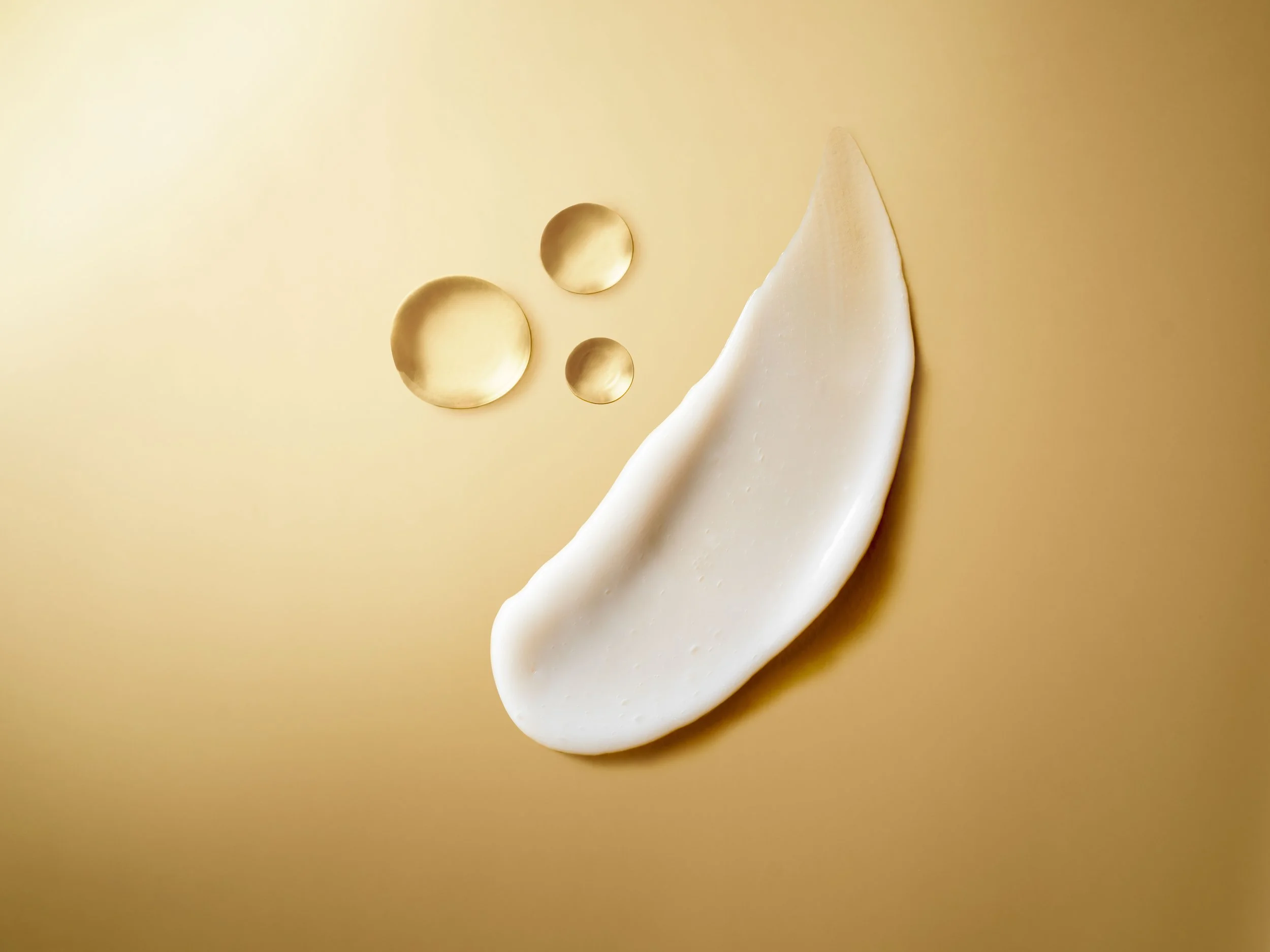White cosmetic cream smear with three golden oil droplets on a beige background