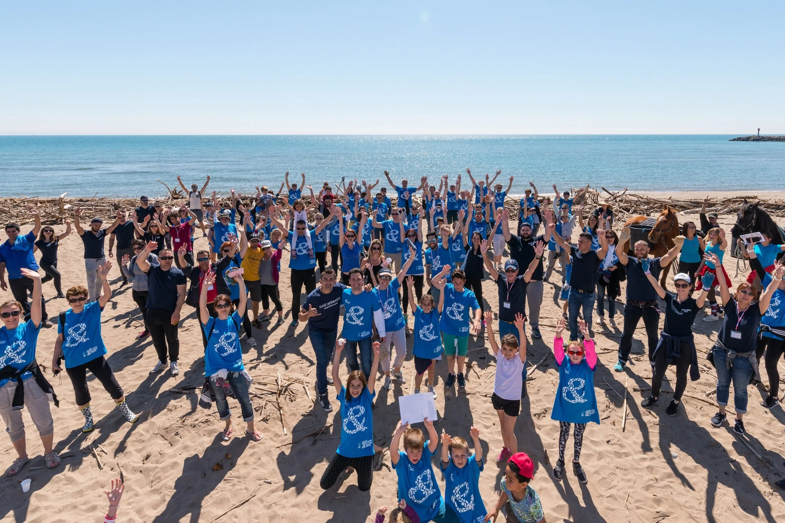 Large group of volunteers in blue shirts celebrating on a driftwood-covered beach beside the ocean