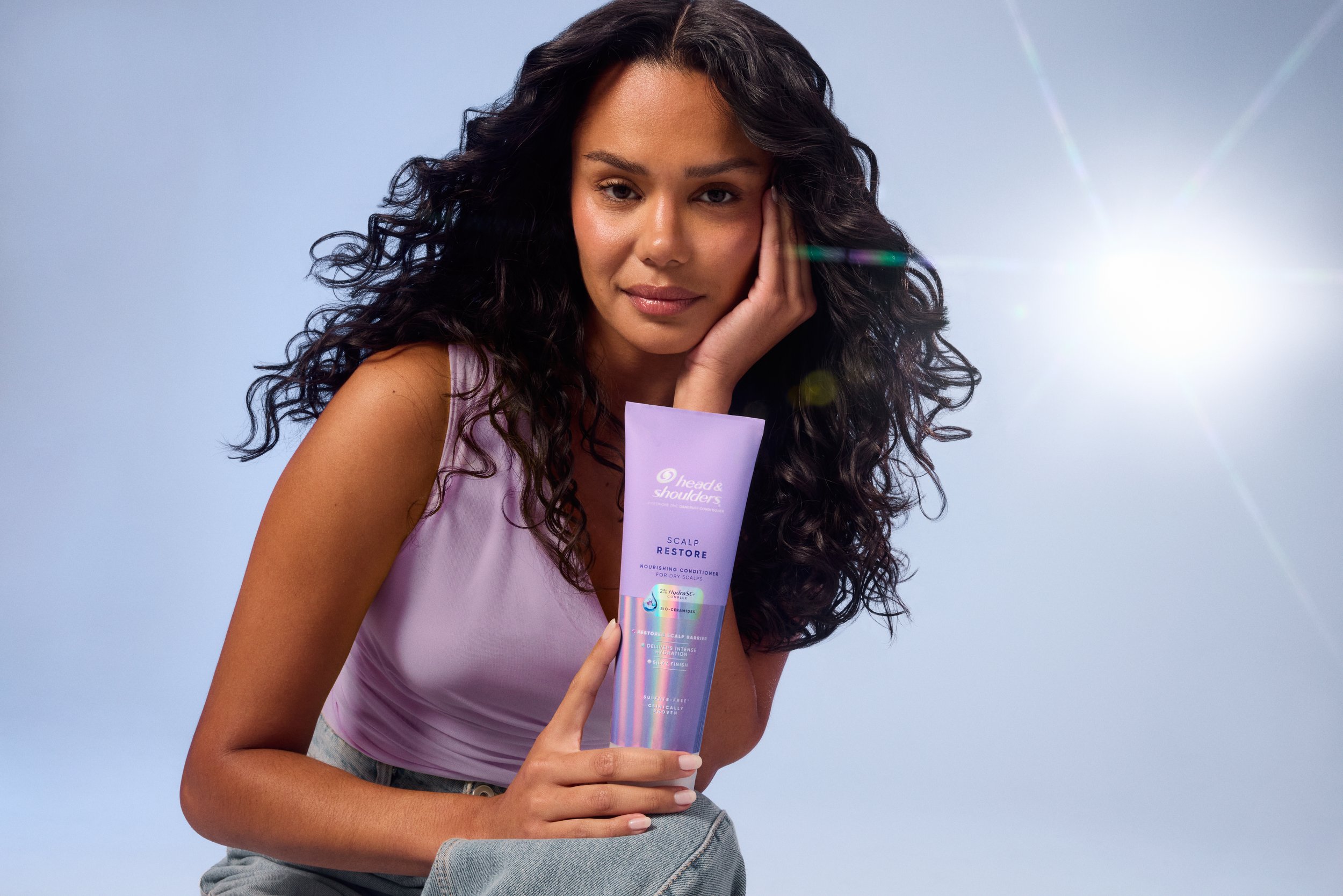 Woman with curly hair holding Head & Shoulders Scalp Restore nourishing conditioner against a blue background