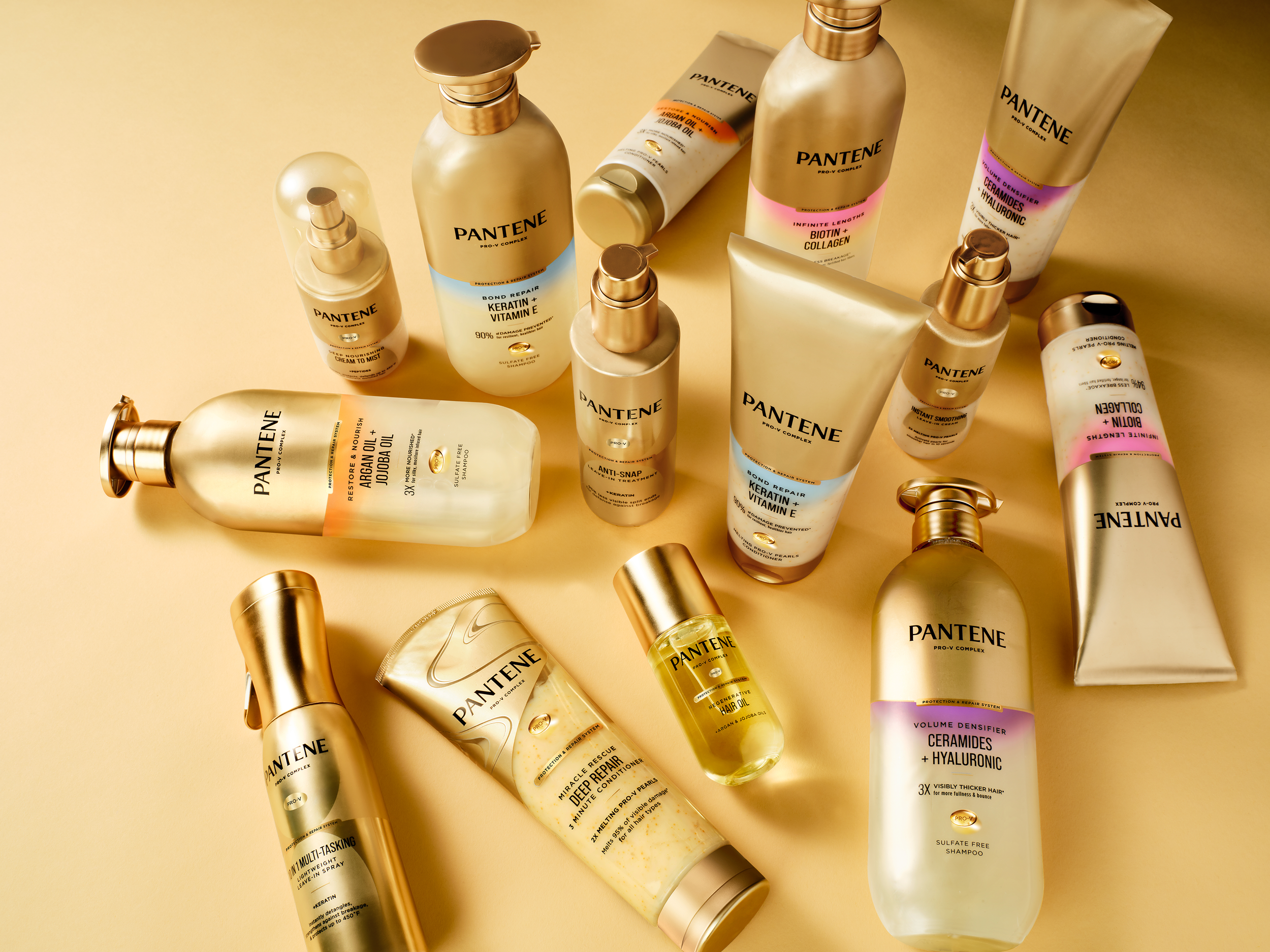 Assortment of Pantene Pro-V hair care products in gold and cream bottles, tubes, and pumps arranged on a warm golden background.