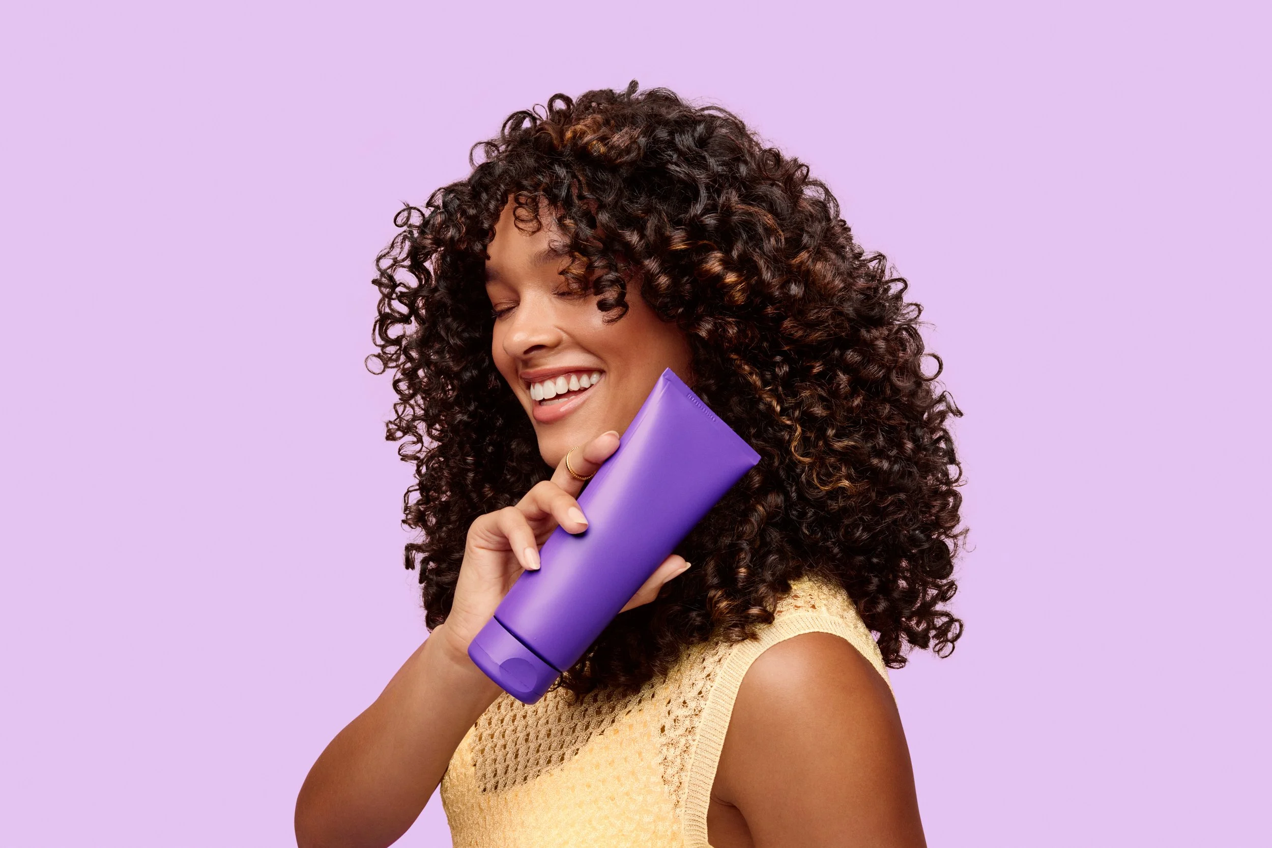 Smiling woman with voluminous curly hair holding a purple hair care product against a lavender background