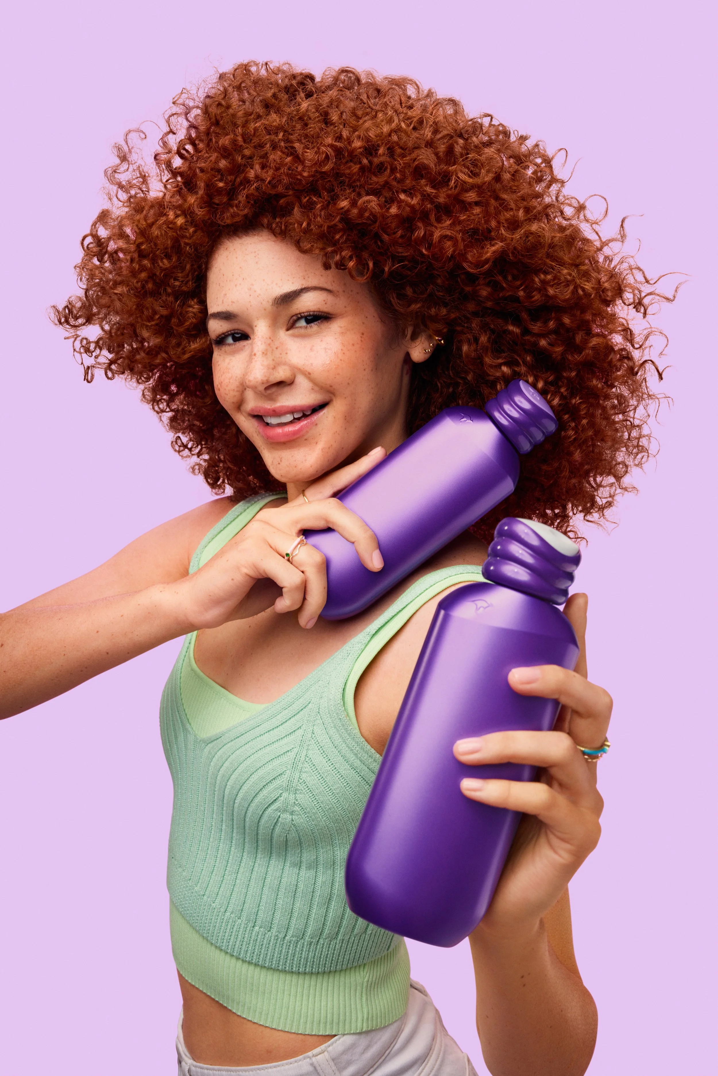 Smiling woman with curly red hair holding purple dumbbells against a lavender background