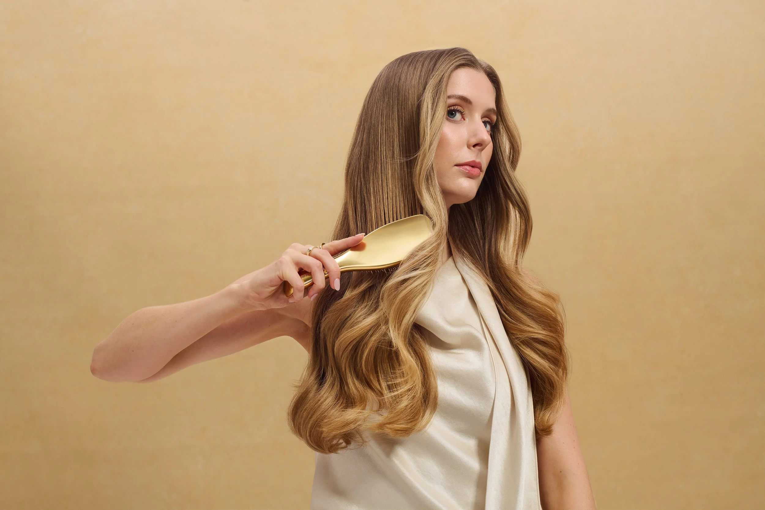 Woman with long wavy blonde hair brushing it with a gold hairbrush against a neutral tan background