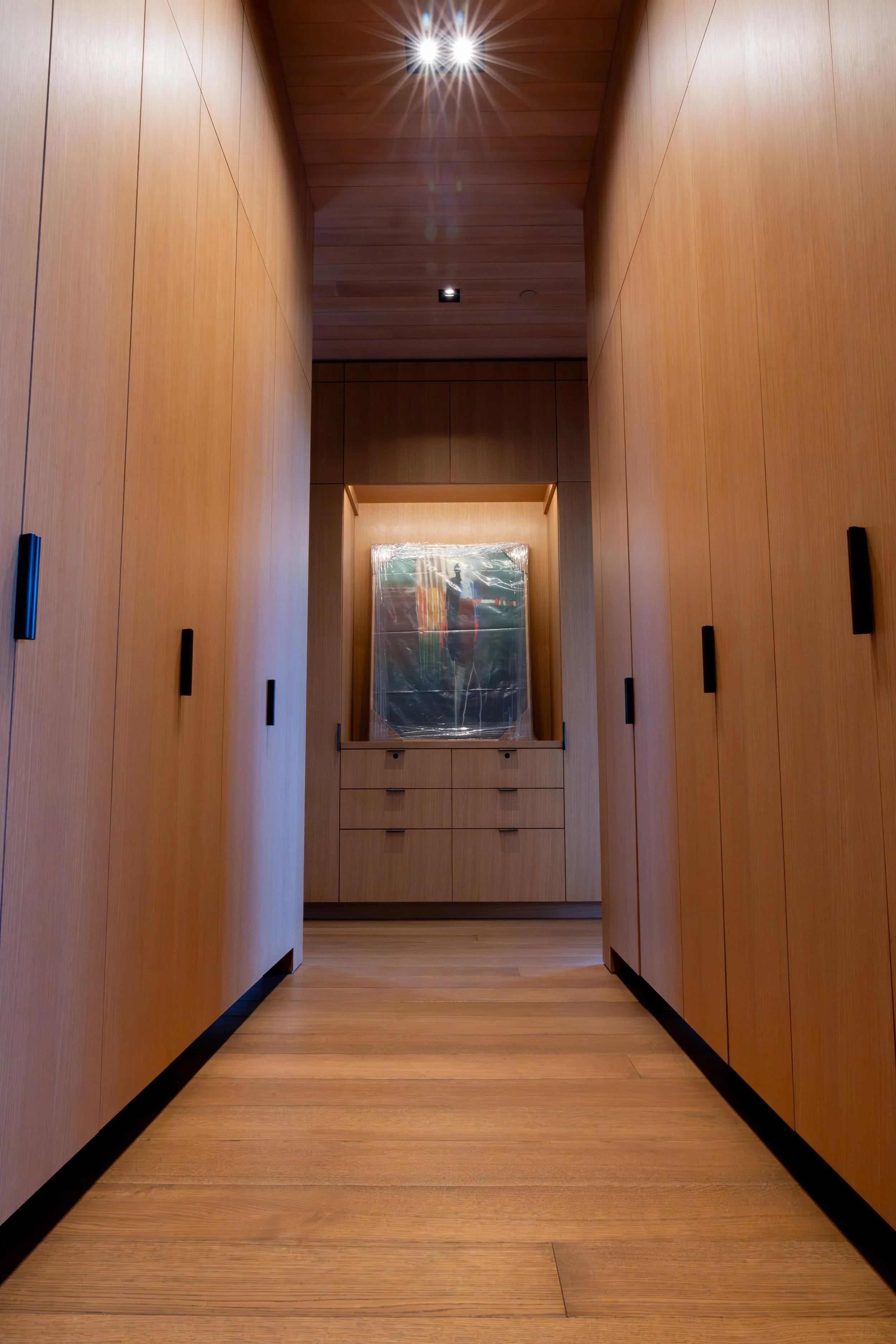 A narrow hallway with wooden walls and ceiling, black handles on the cabinets, and a wooden floor leading to a decorated wall with a framed picture partially covered by plastic.