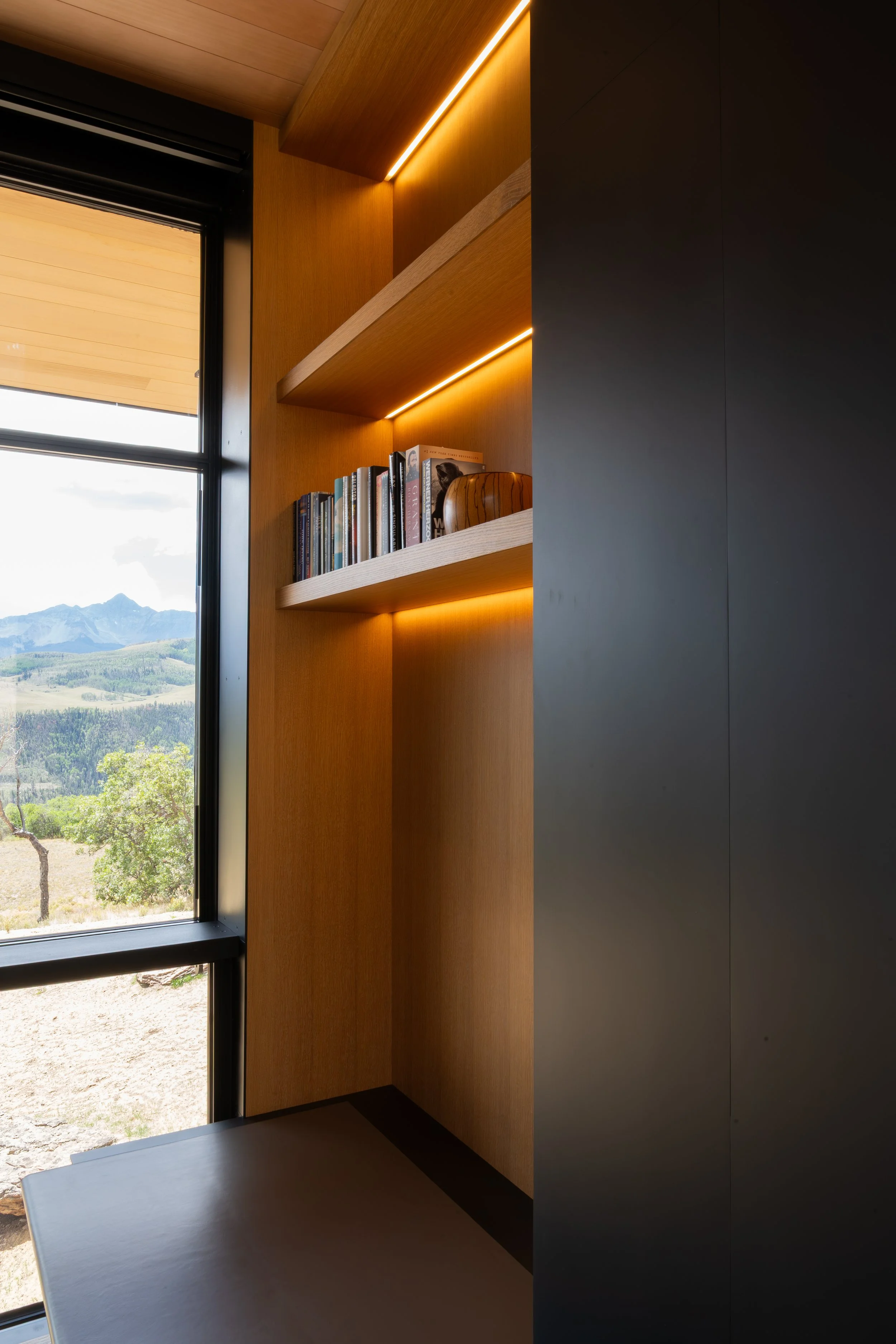 Interior corner of a room with a window showing outdoor landscape, a bookshelf with books, and warm stair lighting on wooden walls.