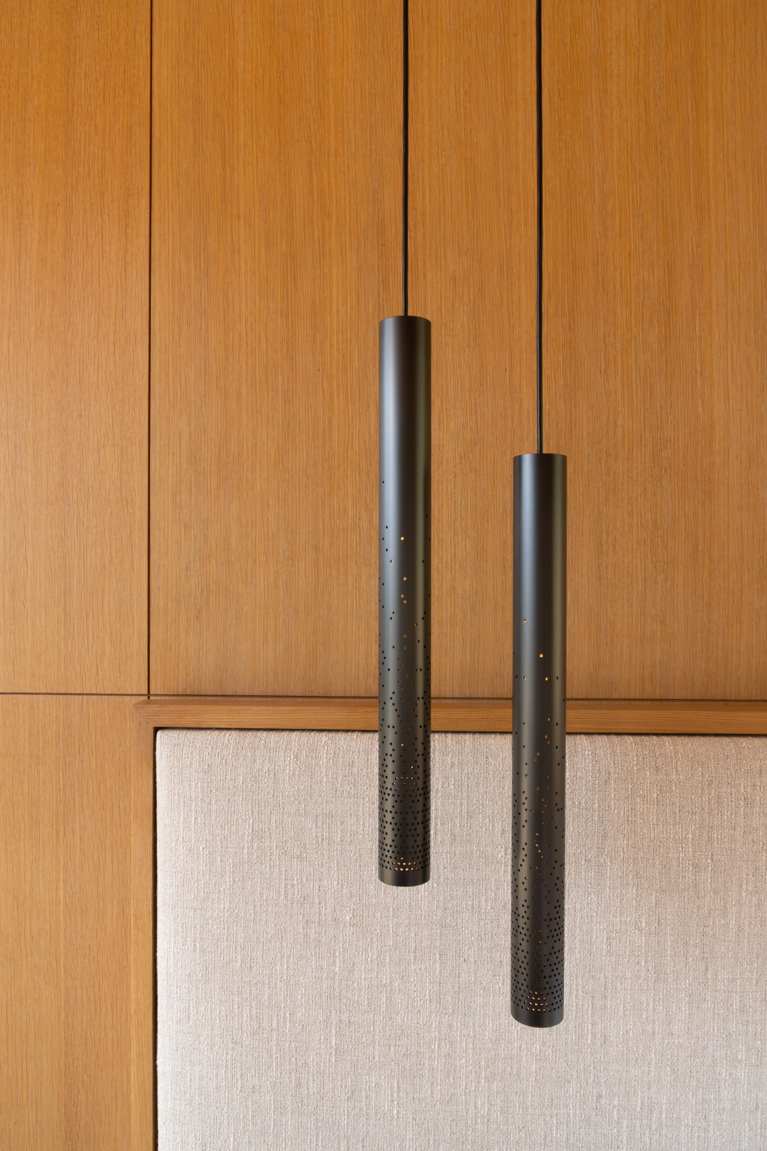 Two modern black pendant lights with perforated ends hanging from black cords, set against a wood-paneled ceiling with a section of beige textured wall.