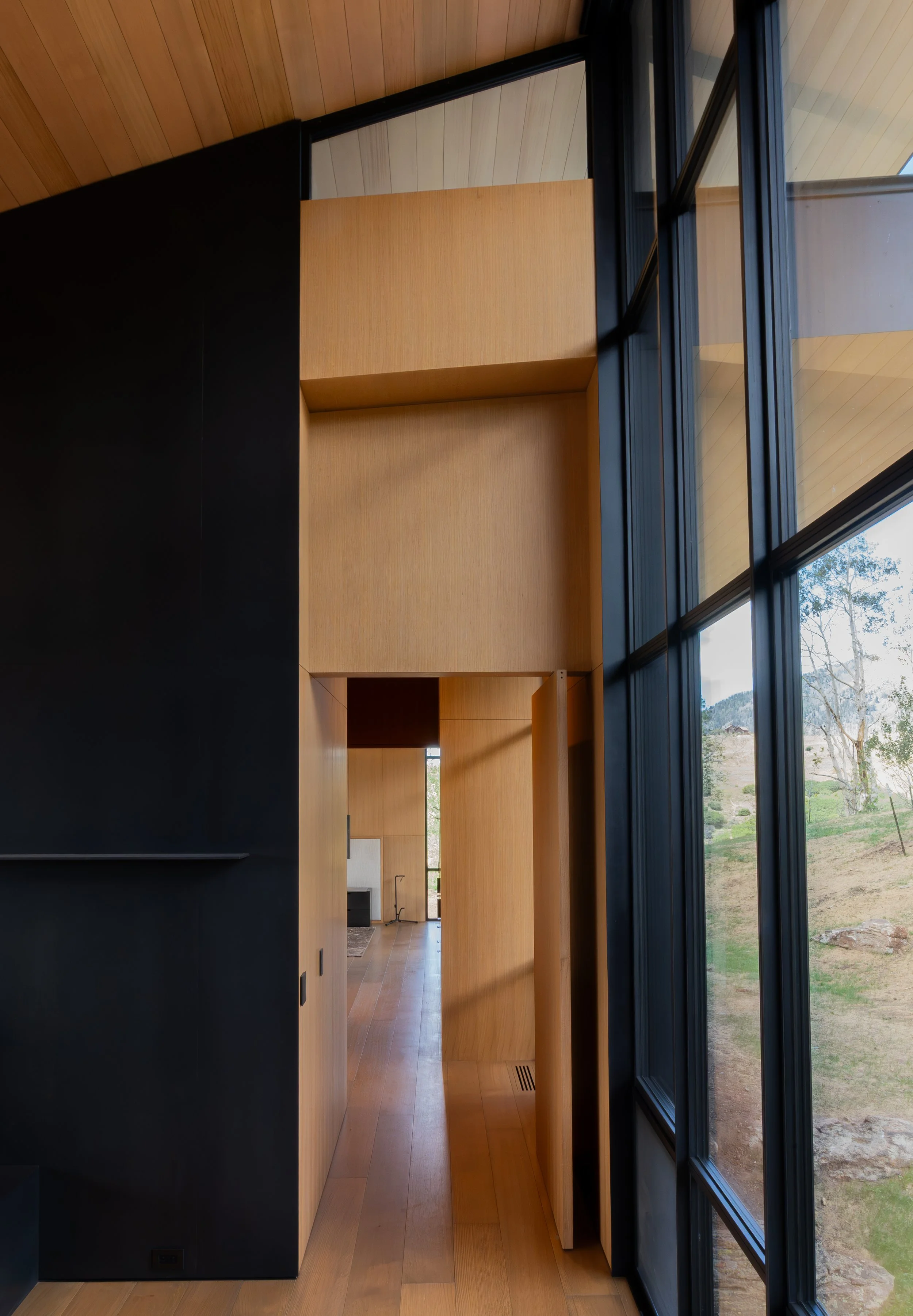 Interior view of a modern house featuring wooden walls, a black wall, and large floor-to-ceiling windows overlooking a landscape with trees and hills.