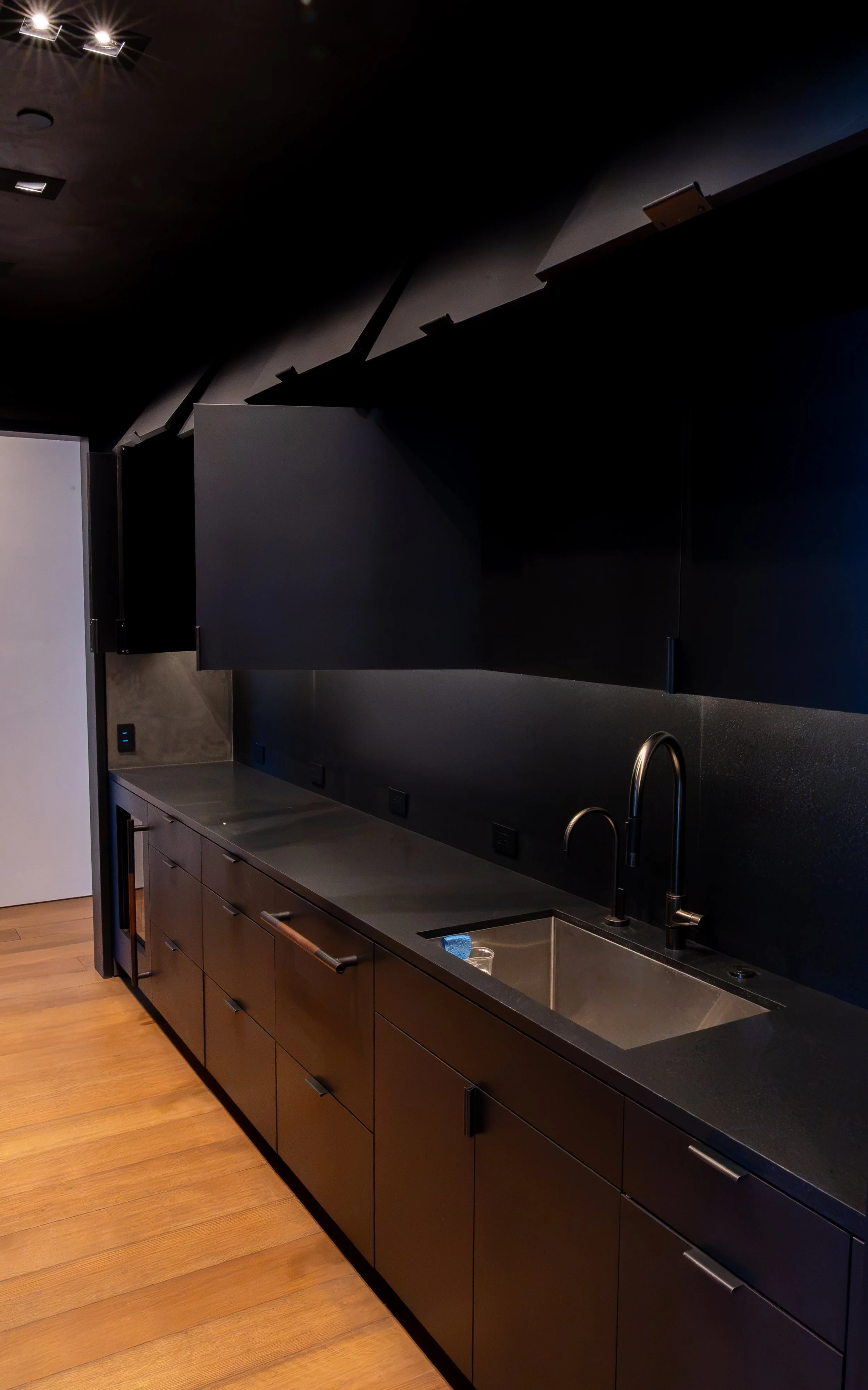Dark modern butlers kitchen with black cabinets, a stainless steel sink, and a black backsplash. Wooden flooring and minimal lighting.