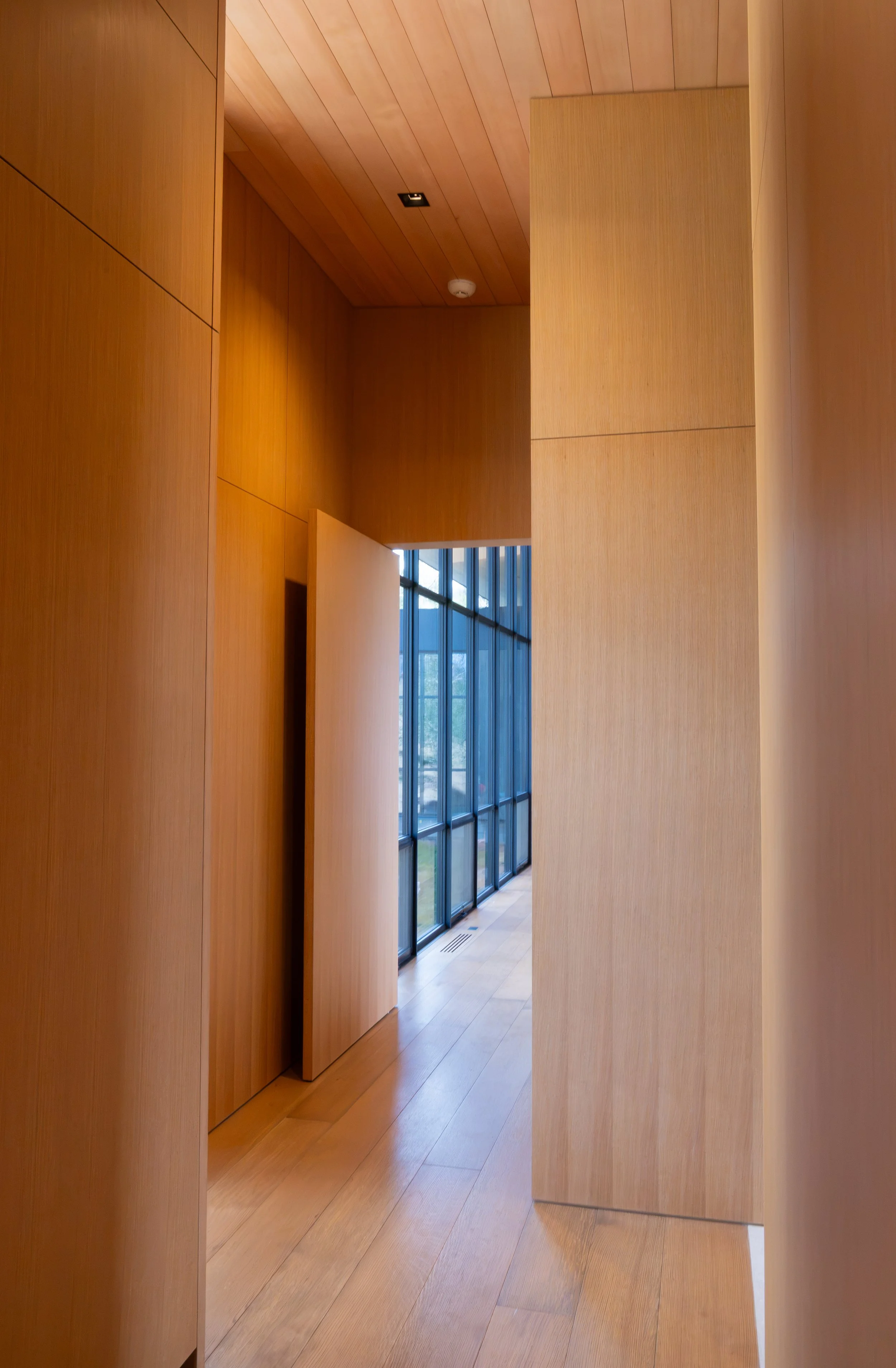 Interior view of a modern home with large windows, wooden floors, and walls, with a view of the outdoors through the windows.