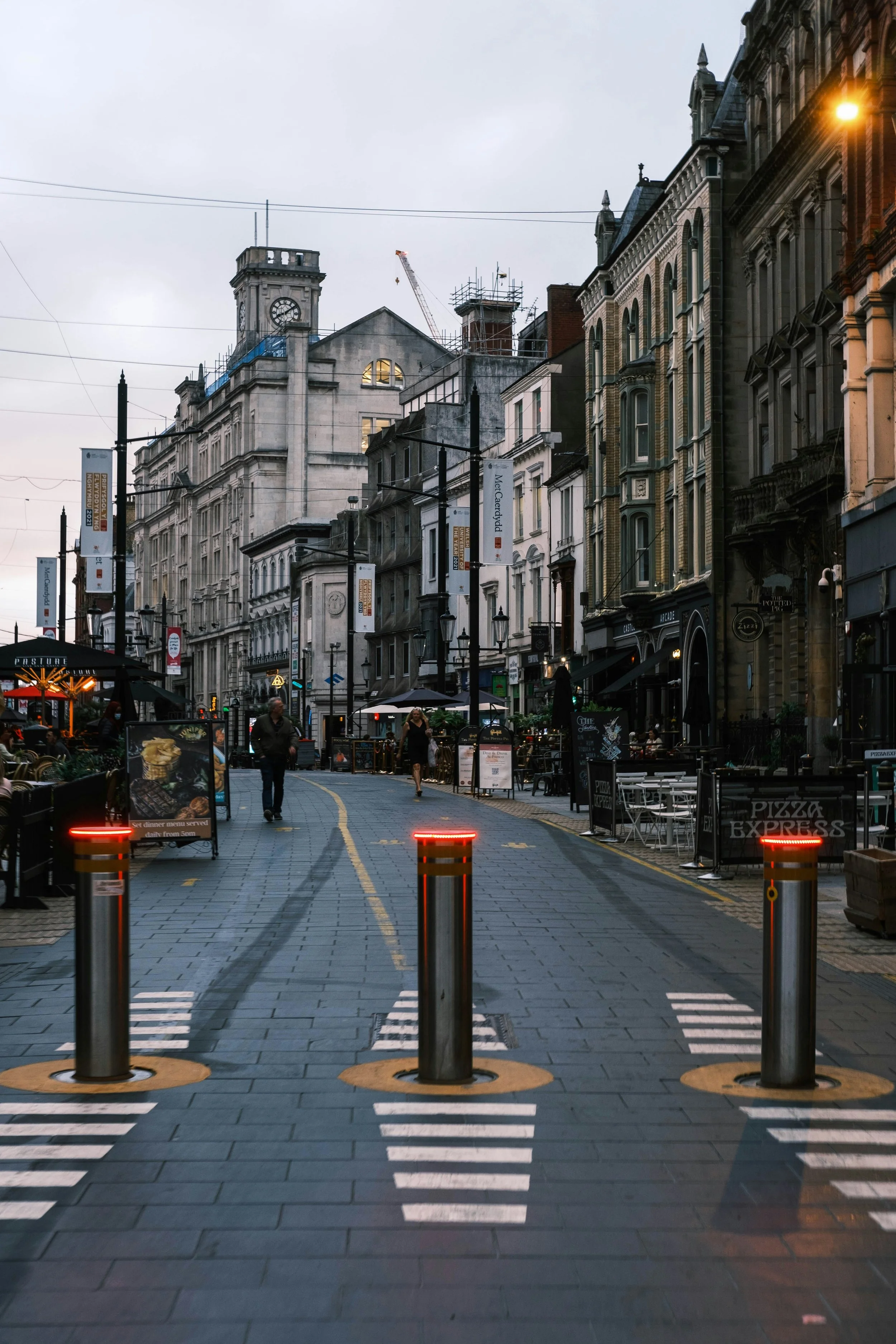 Empty street with bollards, outdoor seating, historical buildings, and a clock tower in the background during dusk.