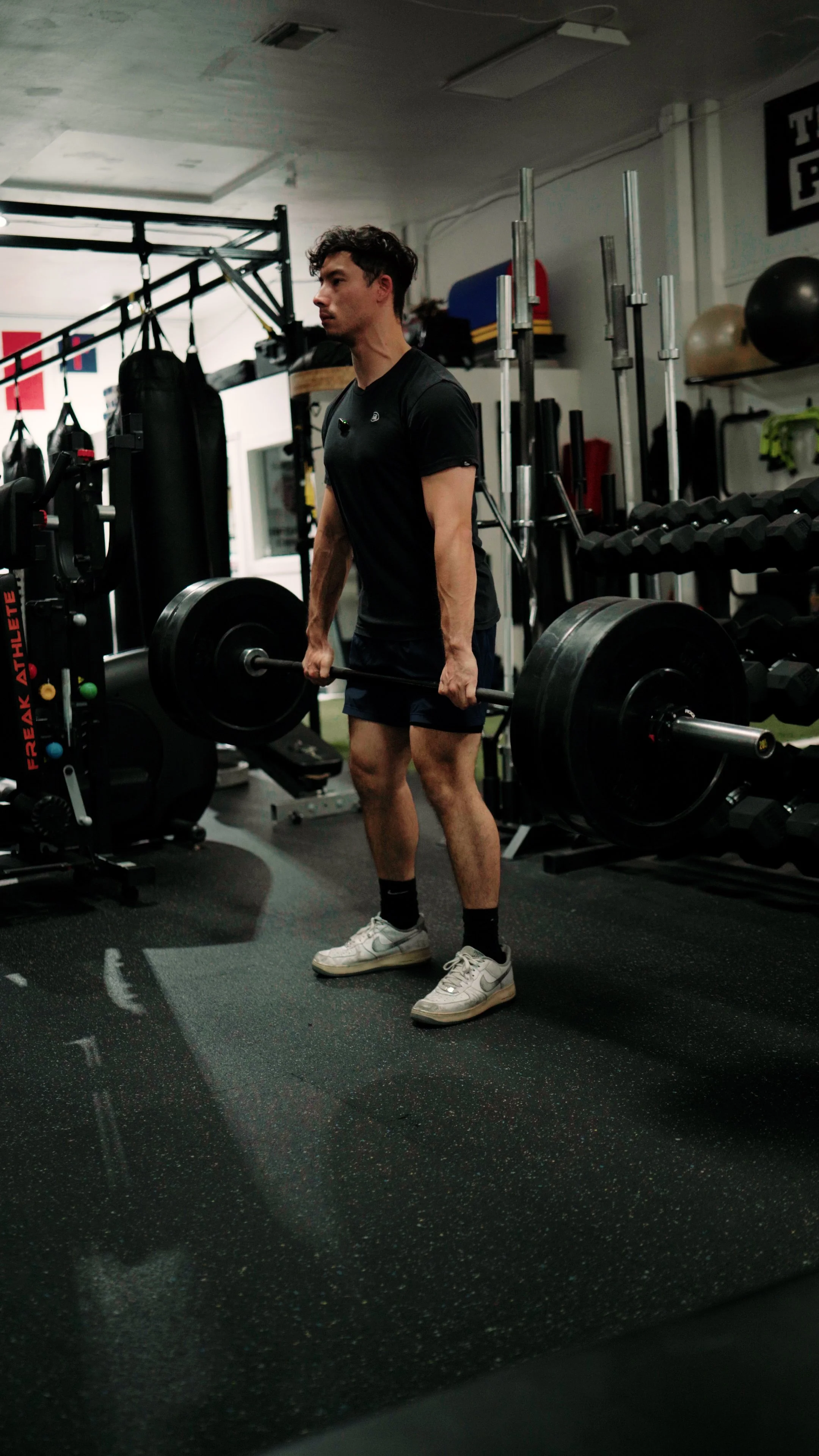 A man in black t-shirt and shorts lifting a barbell in a gym surrounded by various exercise equipment.