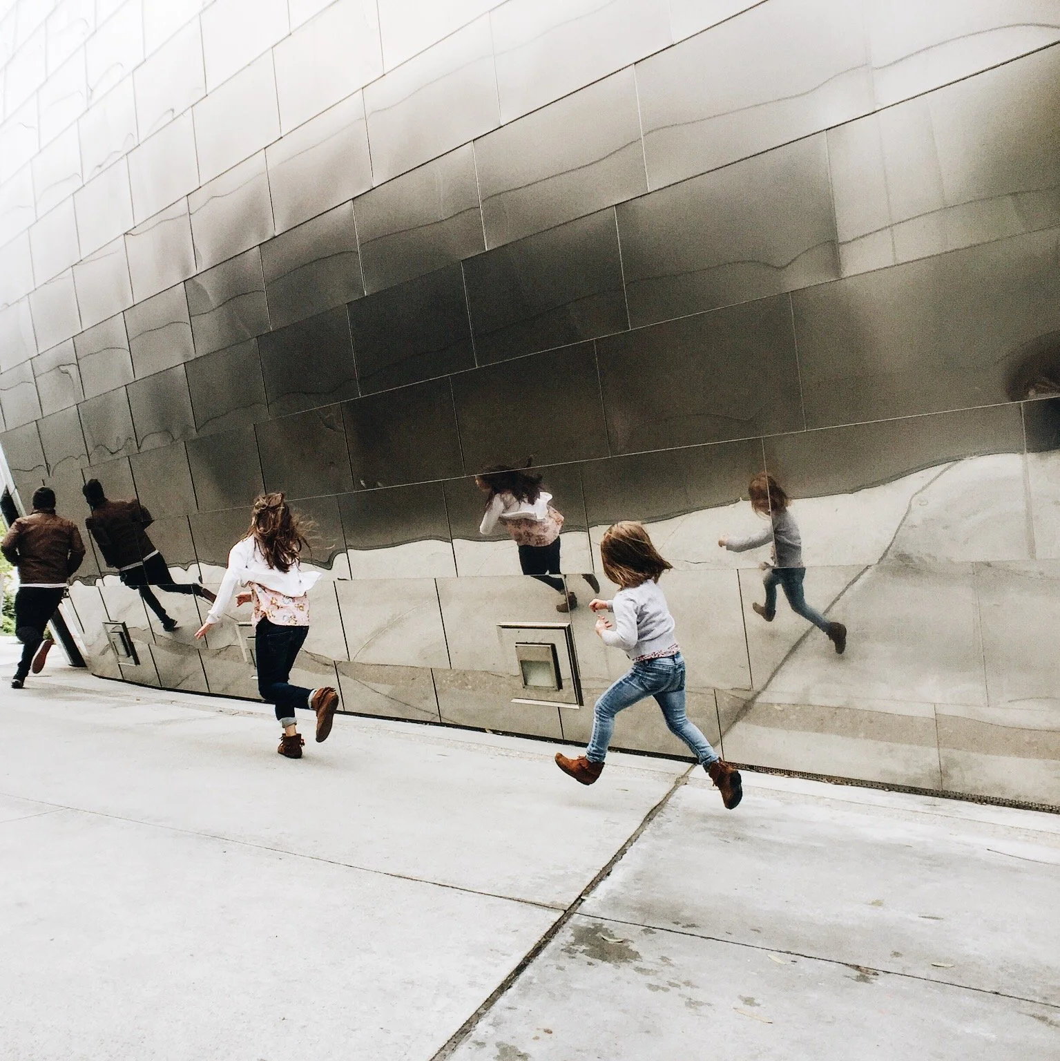 People running outside near a large reflective metallic wall that shows distorted images of them.