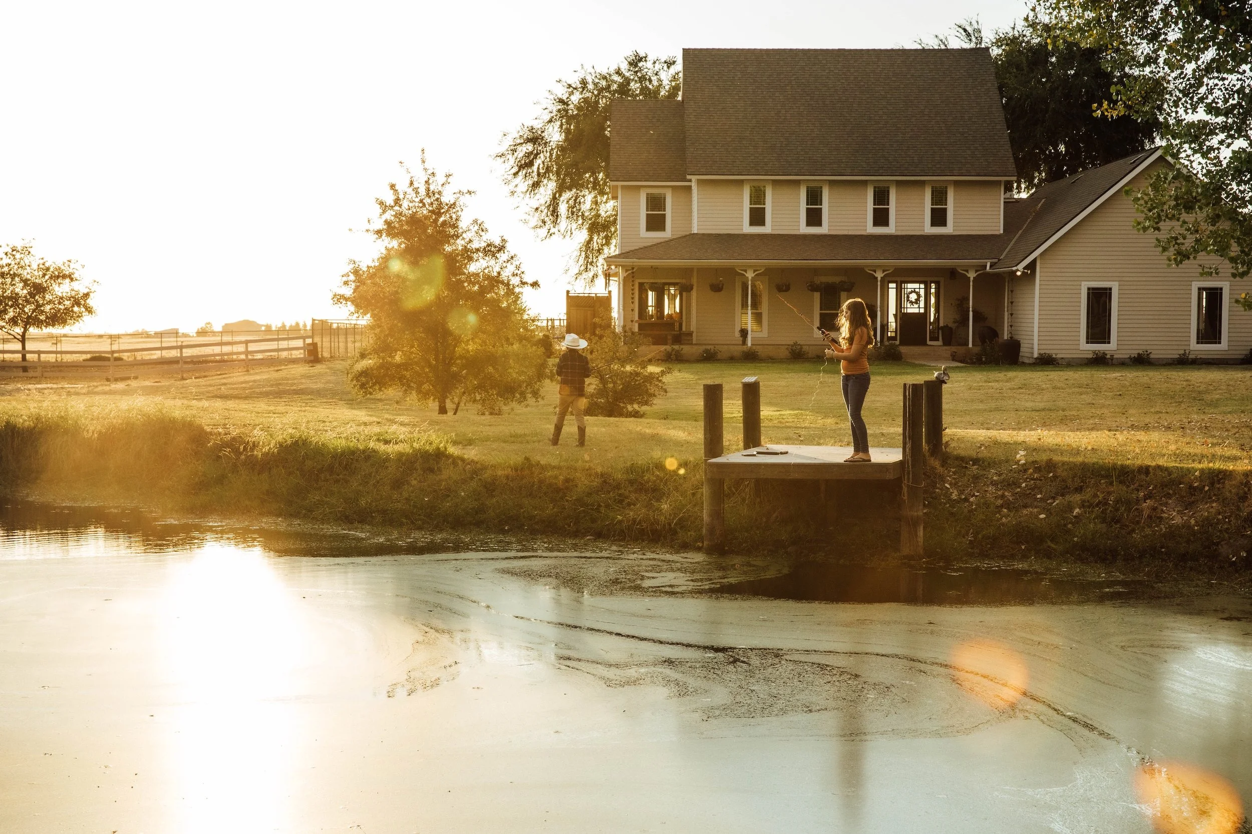 A woman and a child fishing by a pond near a large beige house during sunset, with trees and a fence in the background.