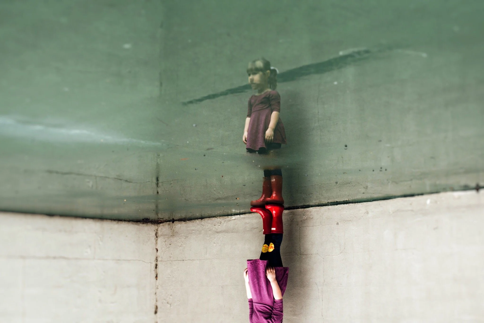 A reflection of a young girl on a handball court, the girl is wearing a purple dress and red rain boots. 