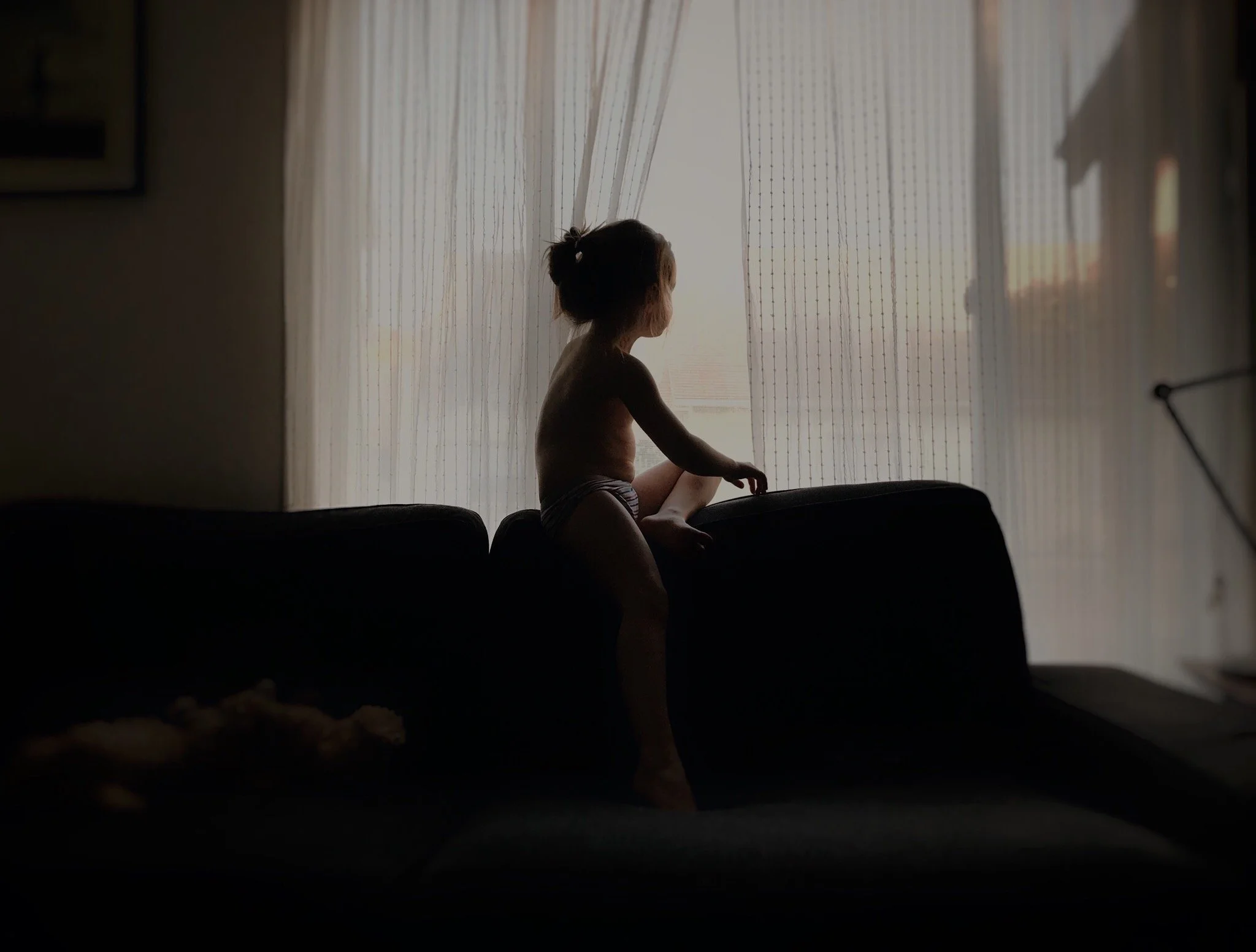 Child sitting on a couch by a window with sheer curtains, silhouette illuminated by daylight.