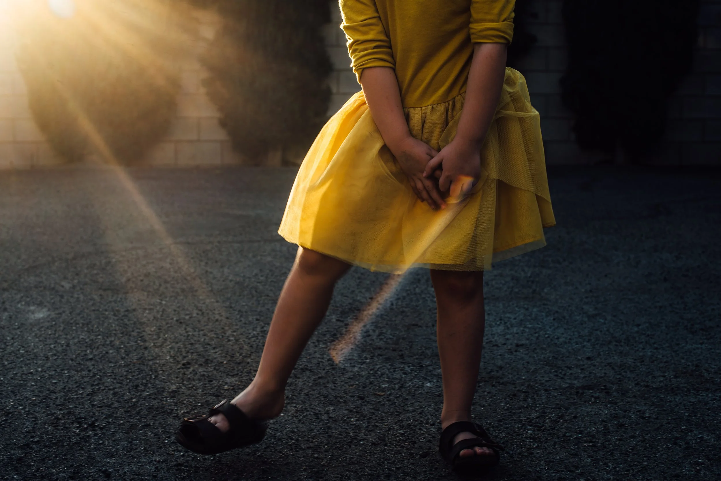 A young girl wearing a yellow dress with tulle fabric, black sandals, standing outdoors on a dark asphalt surface, with her hands clasped in front of her lower abdomen. The background features a brick wall and dark bushes, illuminated by warm sunligh