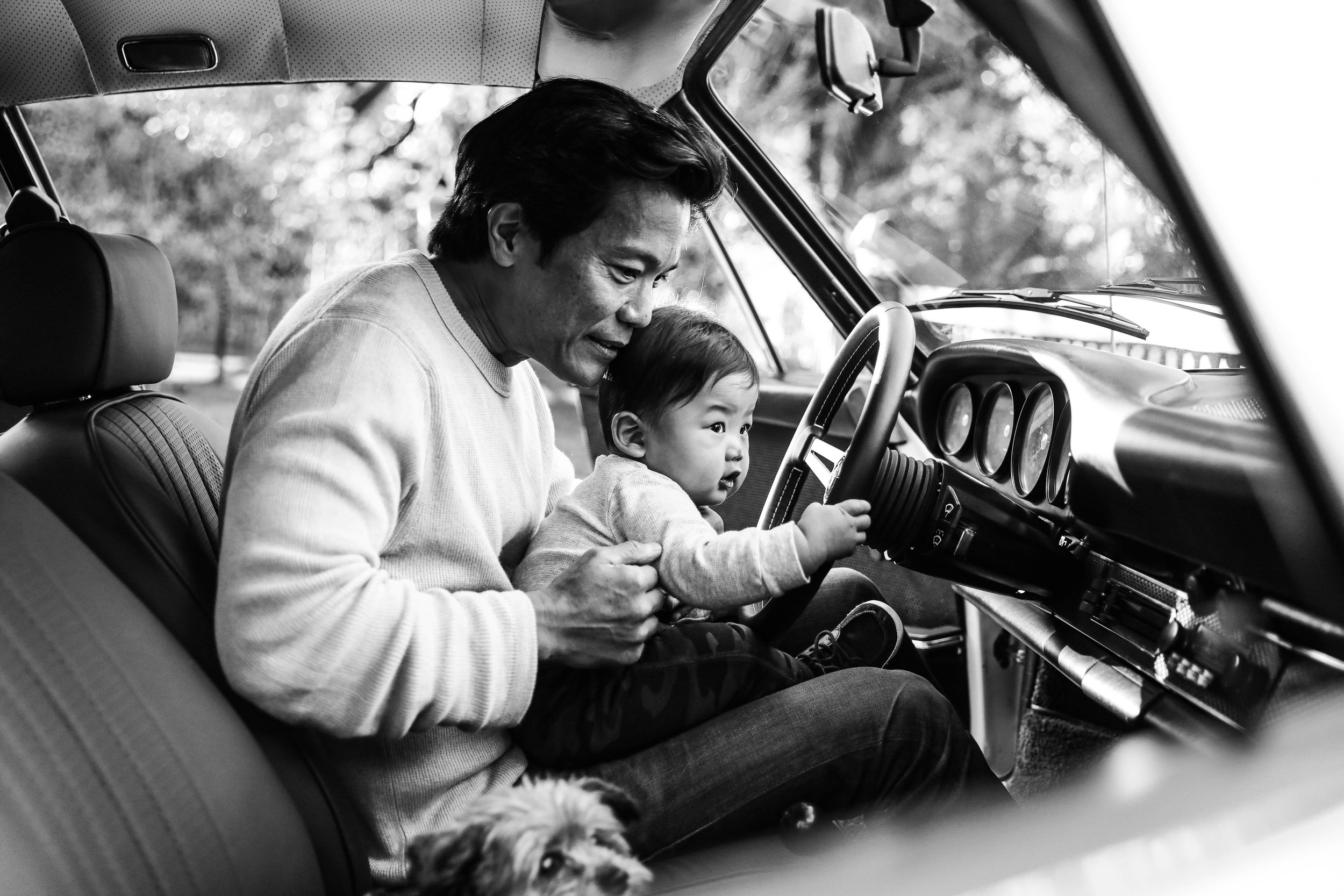 A man and a young child sitting in the front seat of a car, with the man leaning towards the child and the child holding the steering wheel. The scene appears to be outdoors with blurred trees in the background. The image is in black and white.