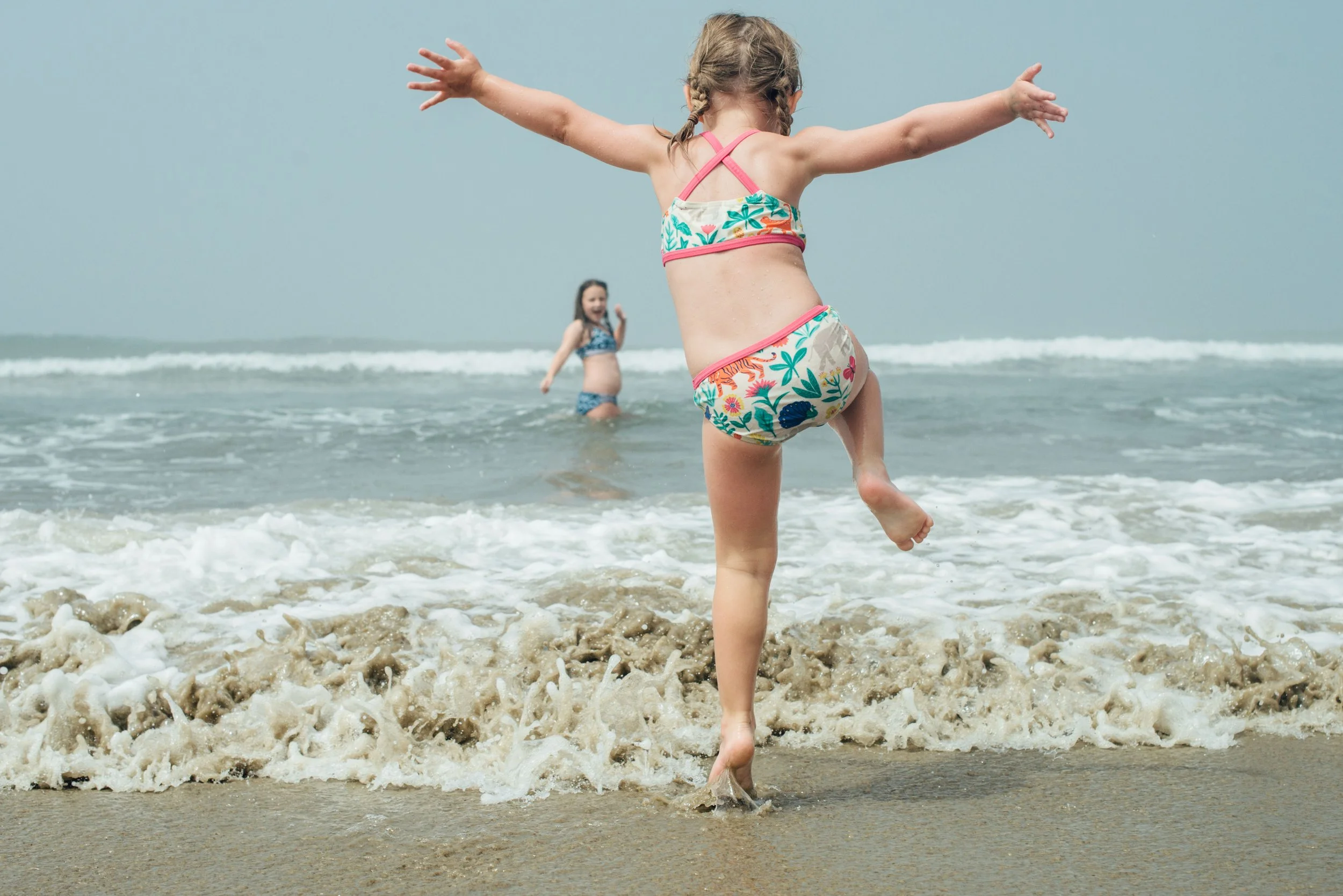 Two young girls playing and running in shallow ocean water near the shoreline, with one girl in the foreground and the other in the background, both wearing swimsuits.
