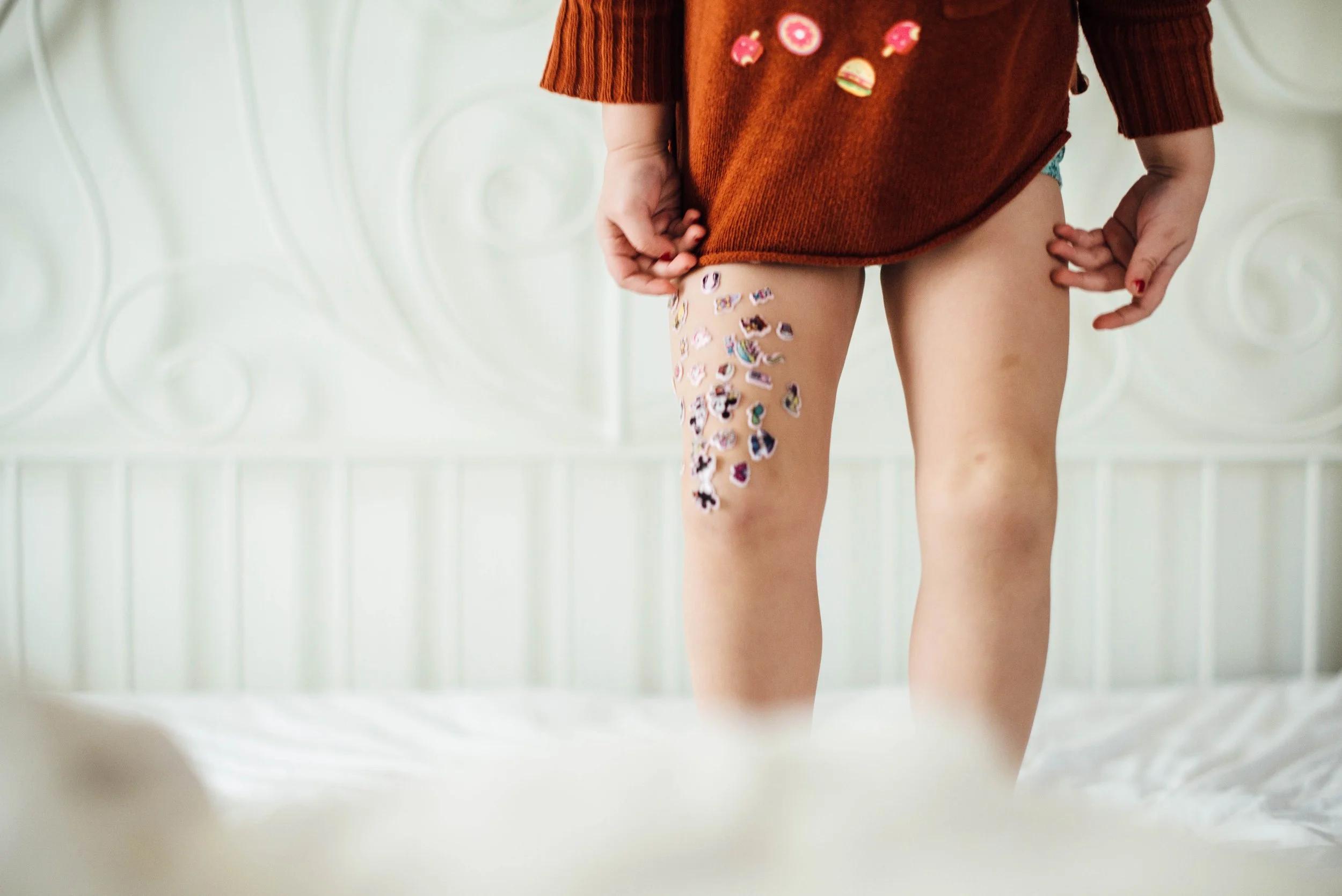 Child pulling up a brown sweater with colorful patches, revealing sticky notes or stickers on their leg, standing against a light-colored, textured headboard.