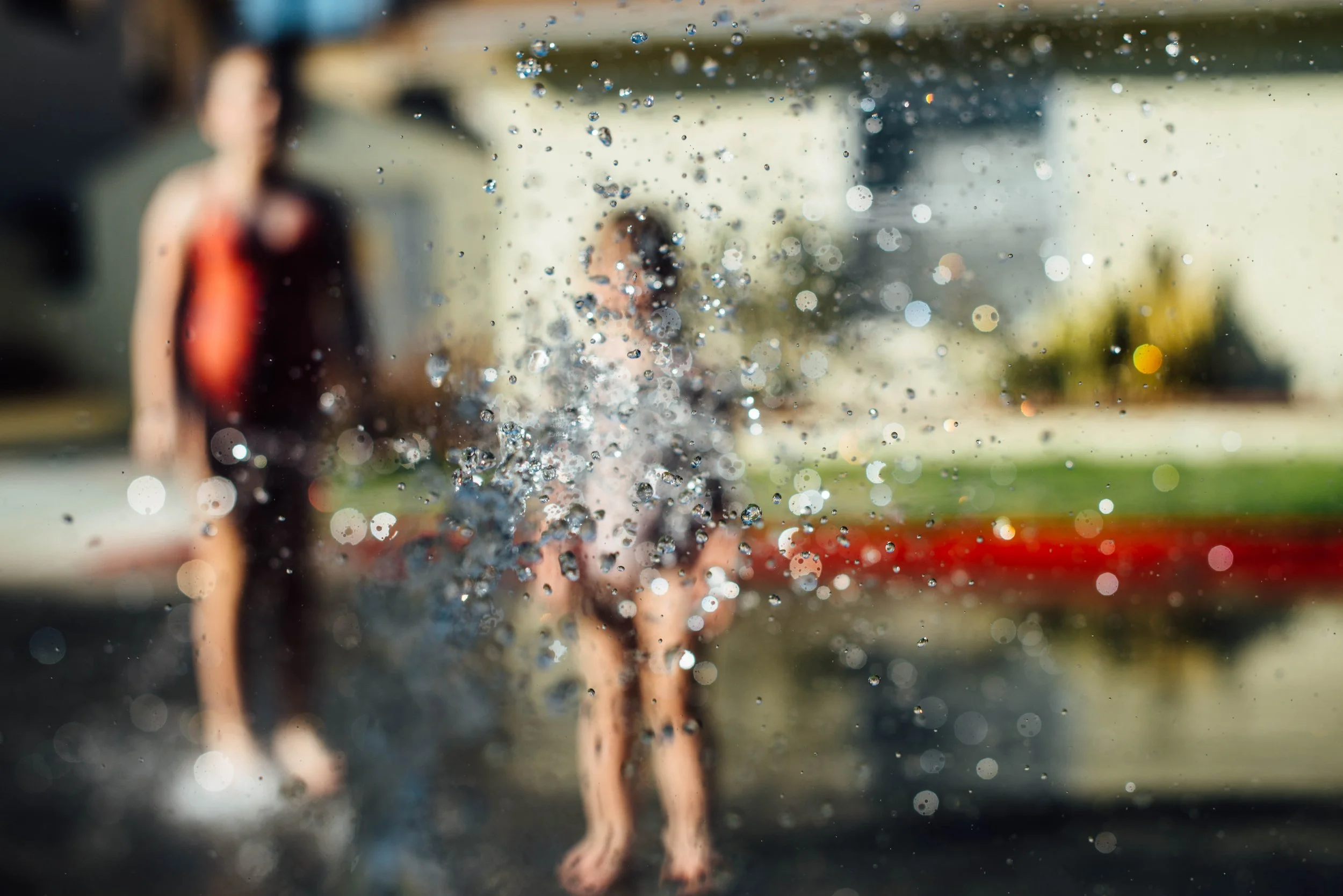 A person is seen through a rain-covered window, with water droplets in focus and the person blurred in the background, standing outside near a house with a lawn.