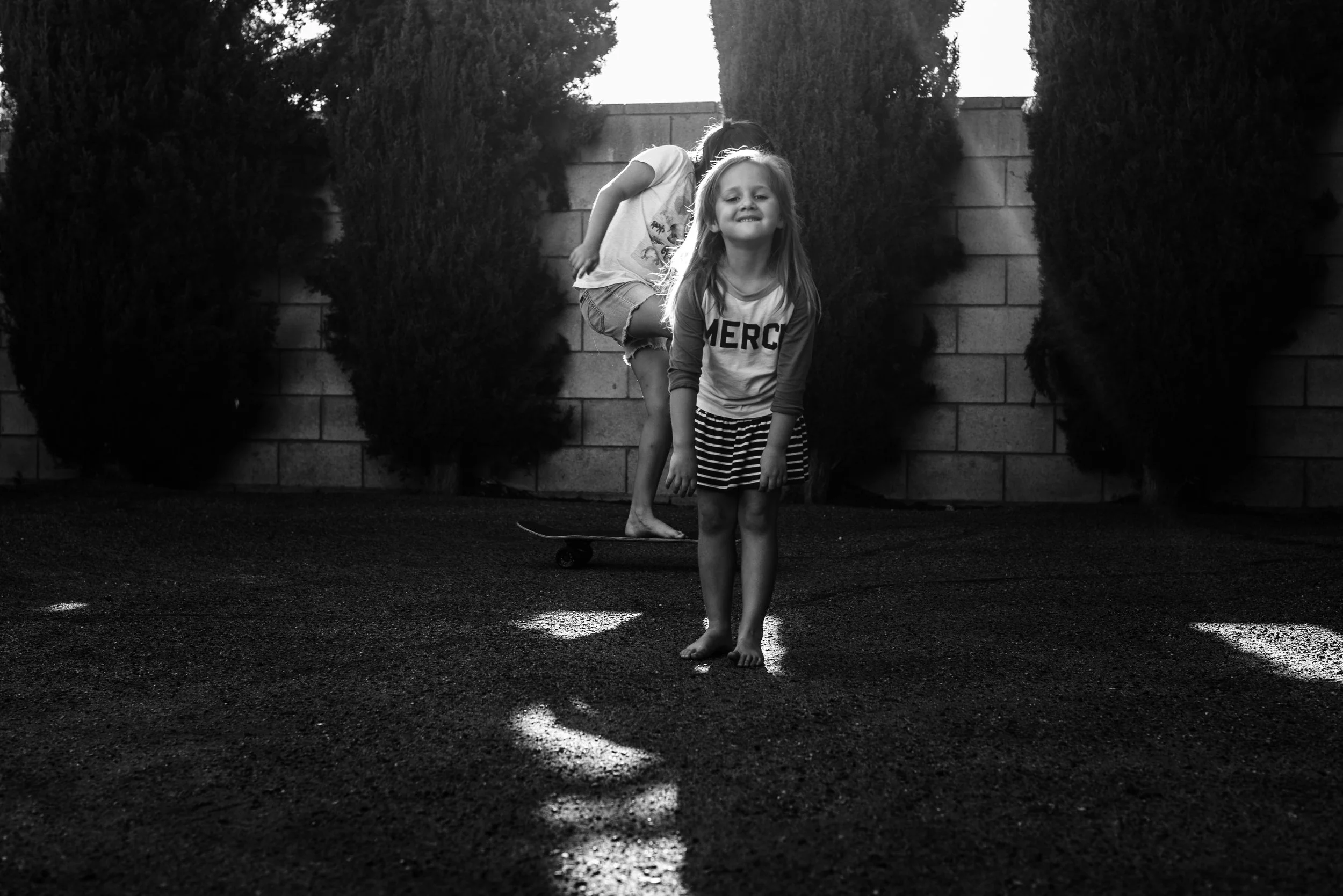 Black and white photo of two young girls outdoors, one in front and one behind, with the girl in front smiling at the camera, standing barefoot, on a textured surface, with tall bushes and a brick wall in the background.