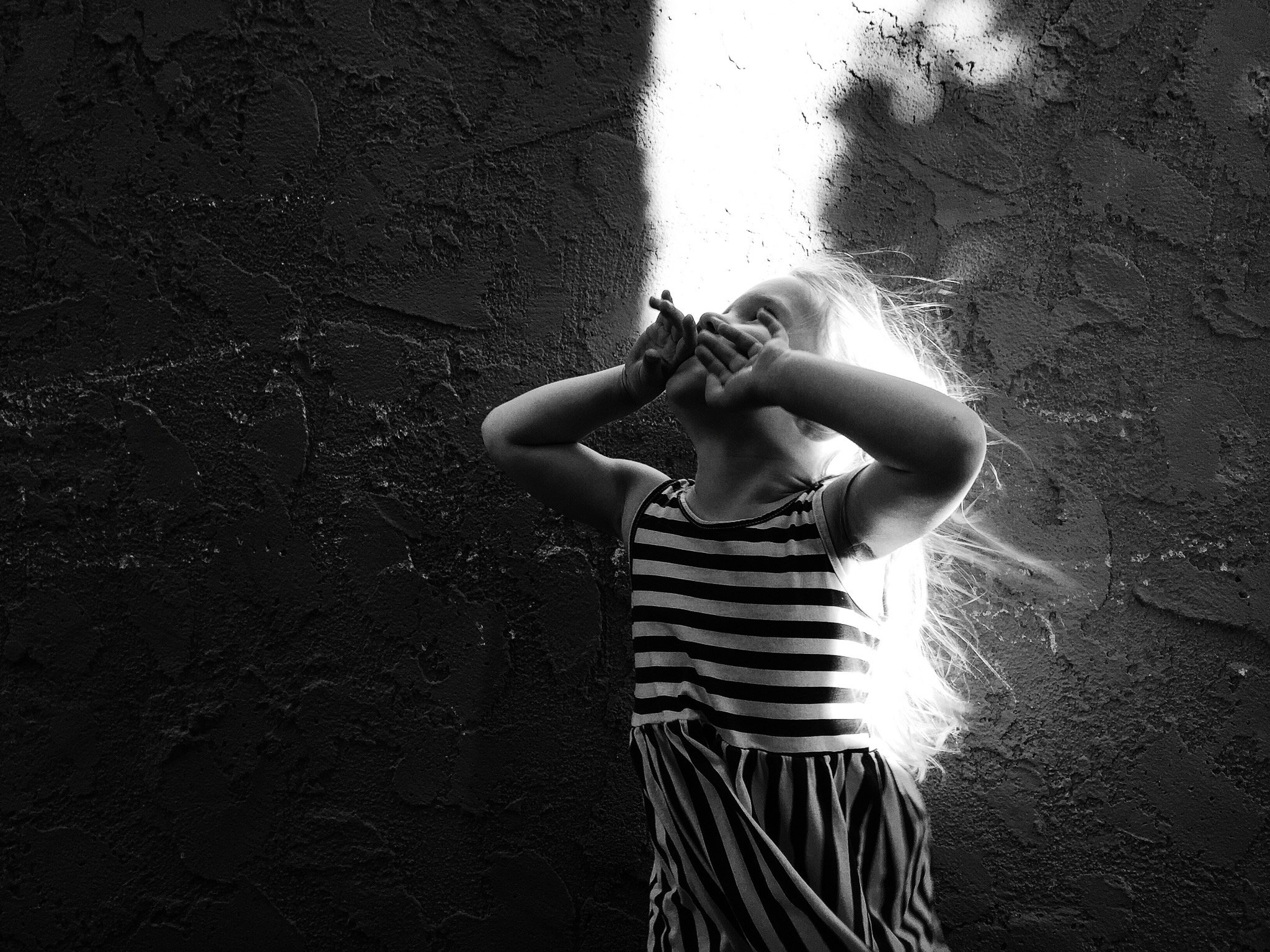 A young girl in a striped shirt is standing against a textured wall, covering part of her face with her hands, with her hair illuminated by light from behind.