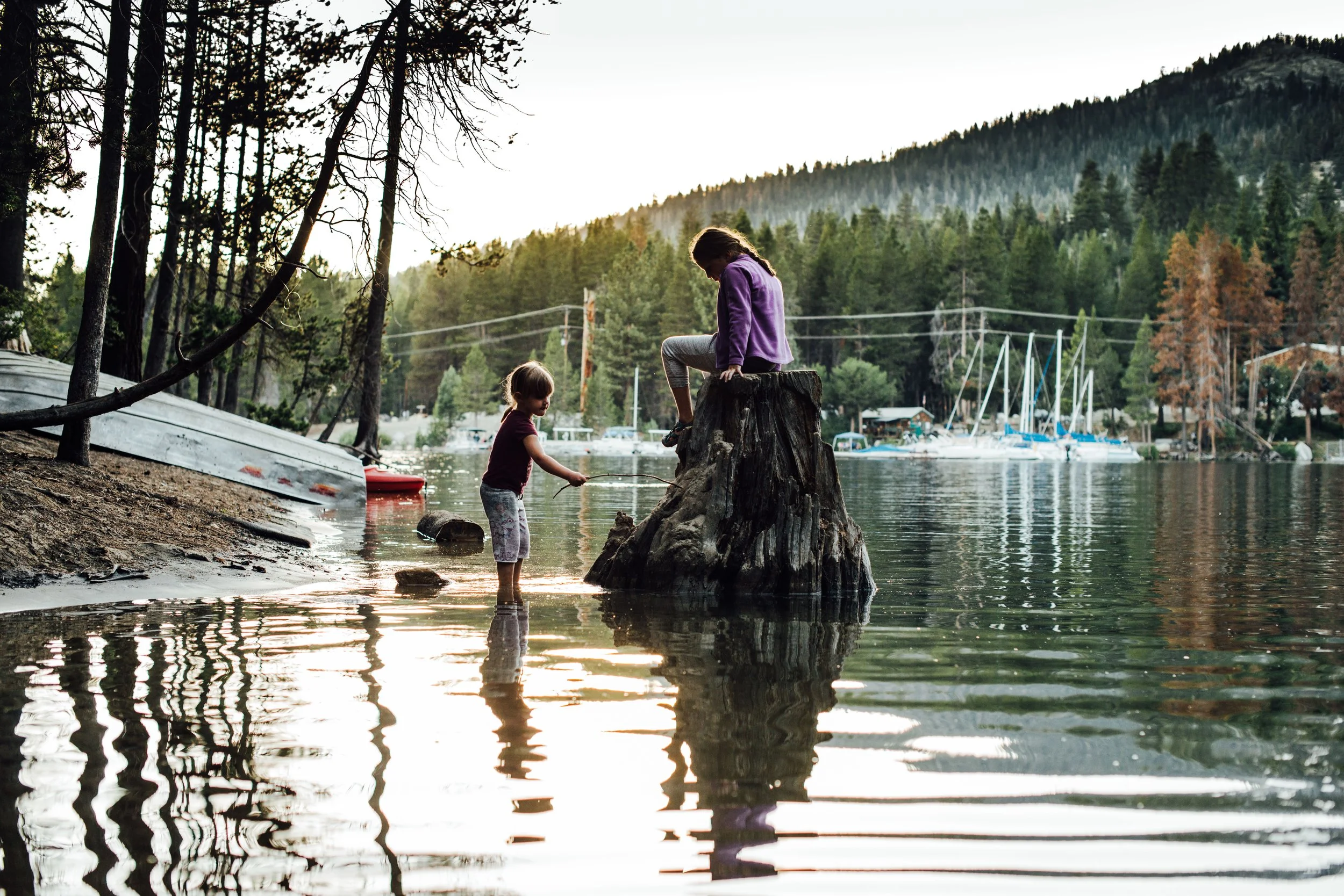 Two children, a girl and a younger boy, are playing in shallow water near a wooded shoreline with boats. The girl is sitting on a tree stump, while the boy is standing in the water, holding a stick. There are trees, boats, and a forested hill in the 